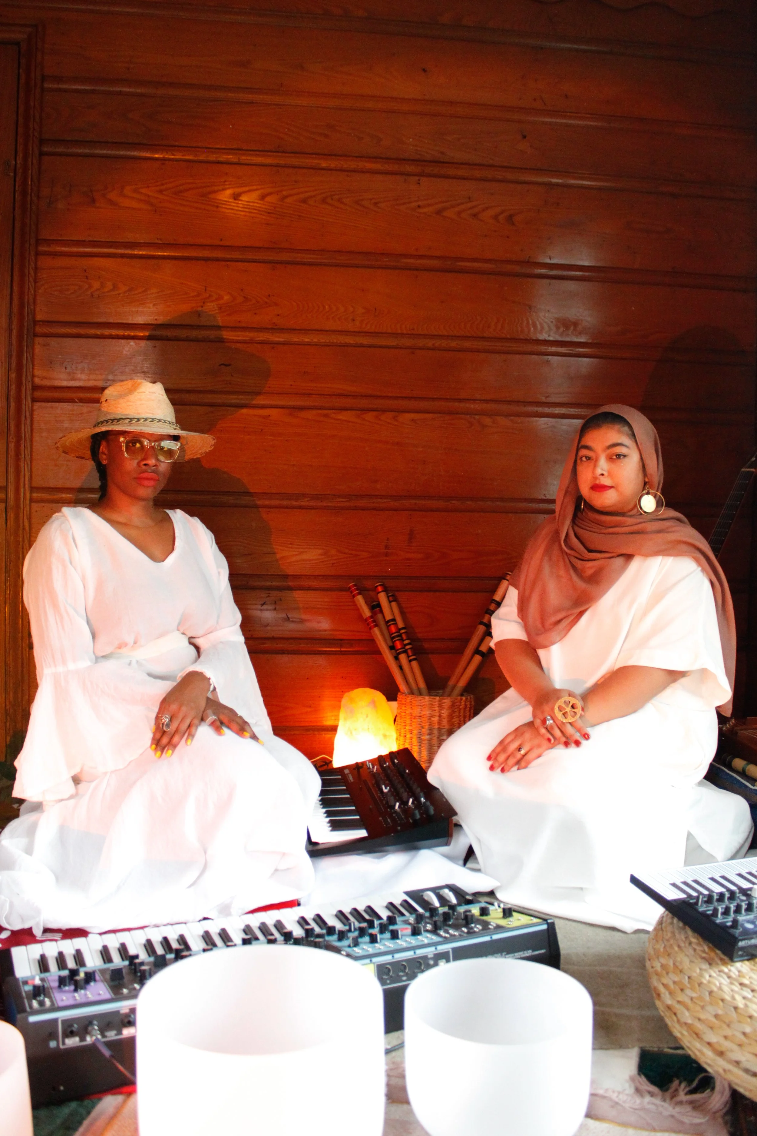Two women sitting on a wooden floor with musical instruments and a salt lamp, dressed in white, with one wearing a hat and sunglasses and the other wearing a scarf and earrings, in a cozy, warm-toned room.