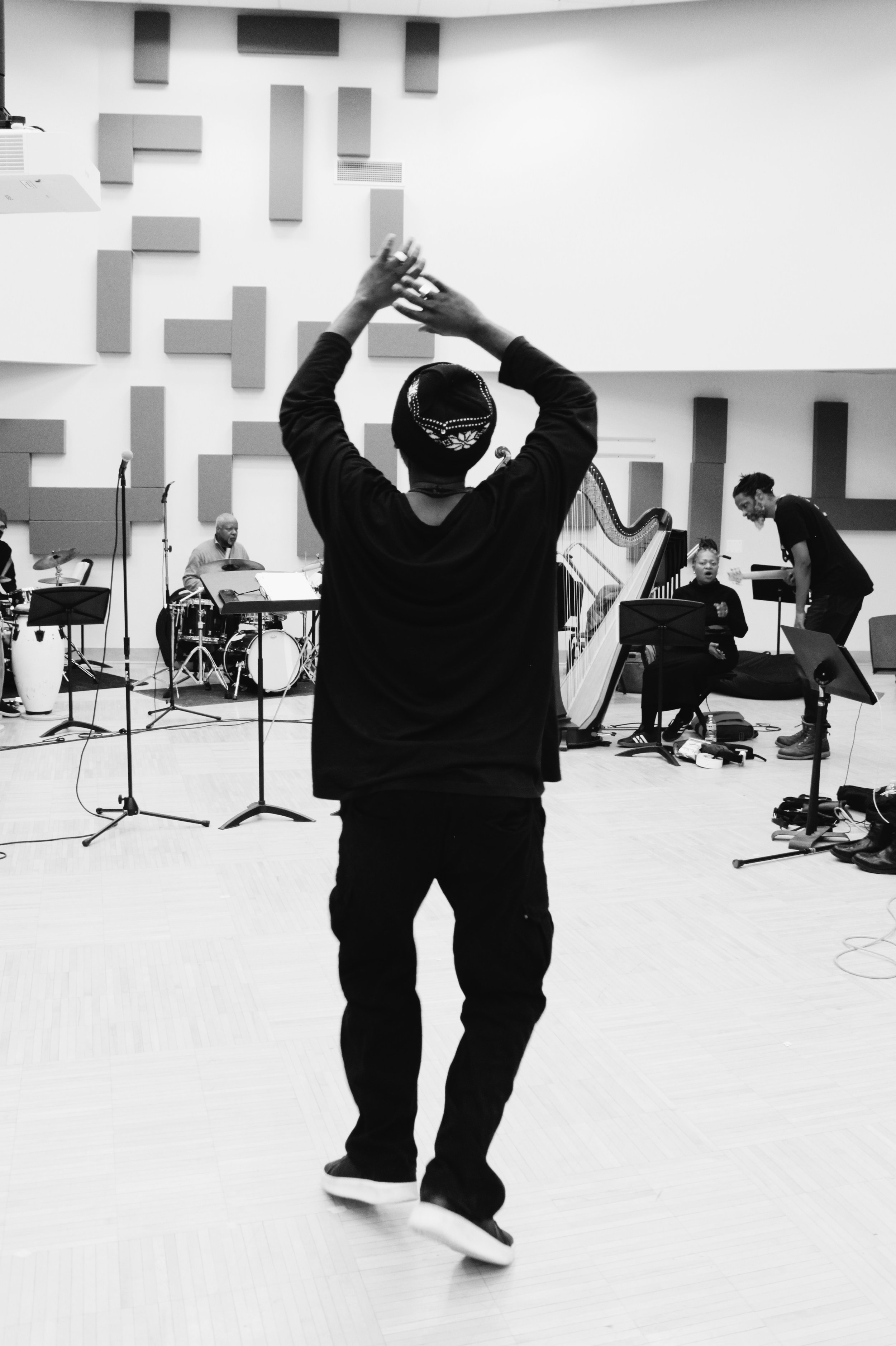 A person stands with arms raised, facing a music ensemble with various instruments including drums, a harp, and a keyboard, in a recording studio or rehearsal space.