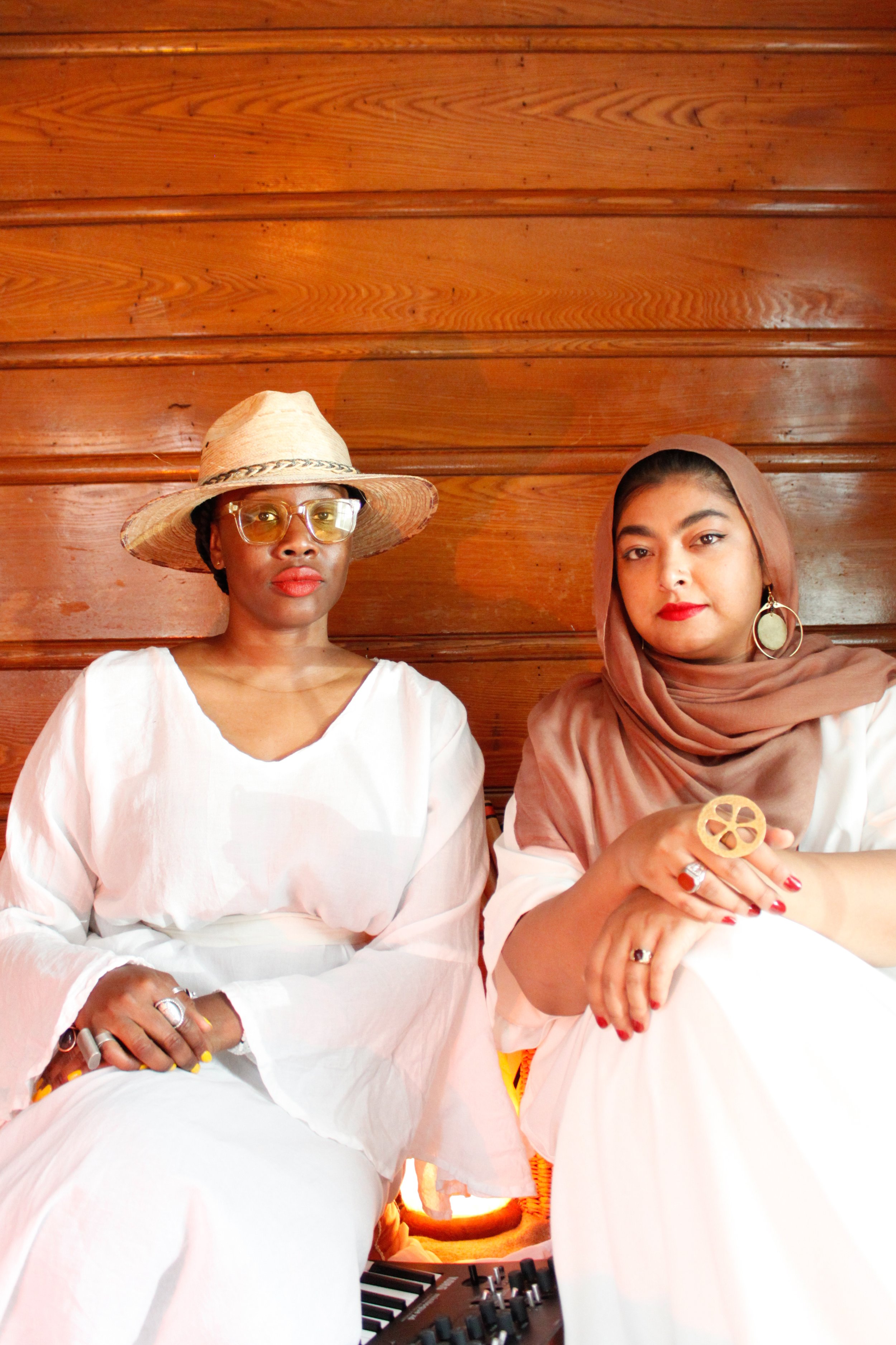 Two women sitting against a wooden wall, dressed in white, one wearing a wide-brimmed hat and yellow-tinted glasses, the other wearing a brown headscarf, with jewelry on their hands and earrings.