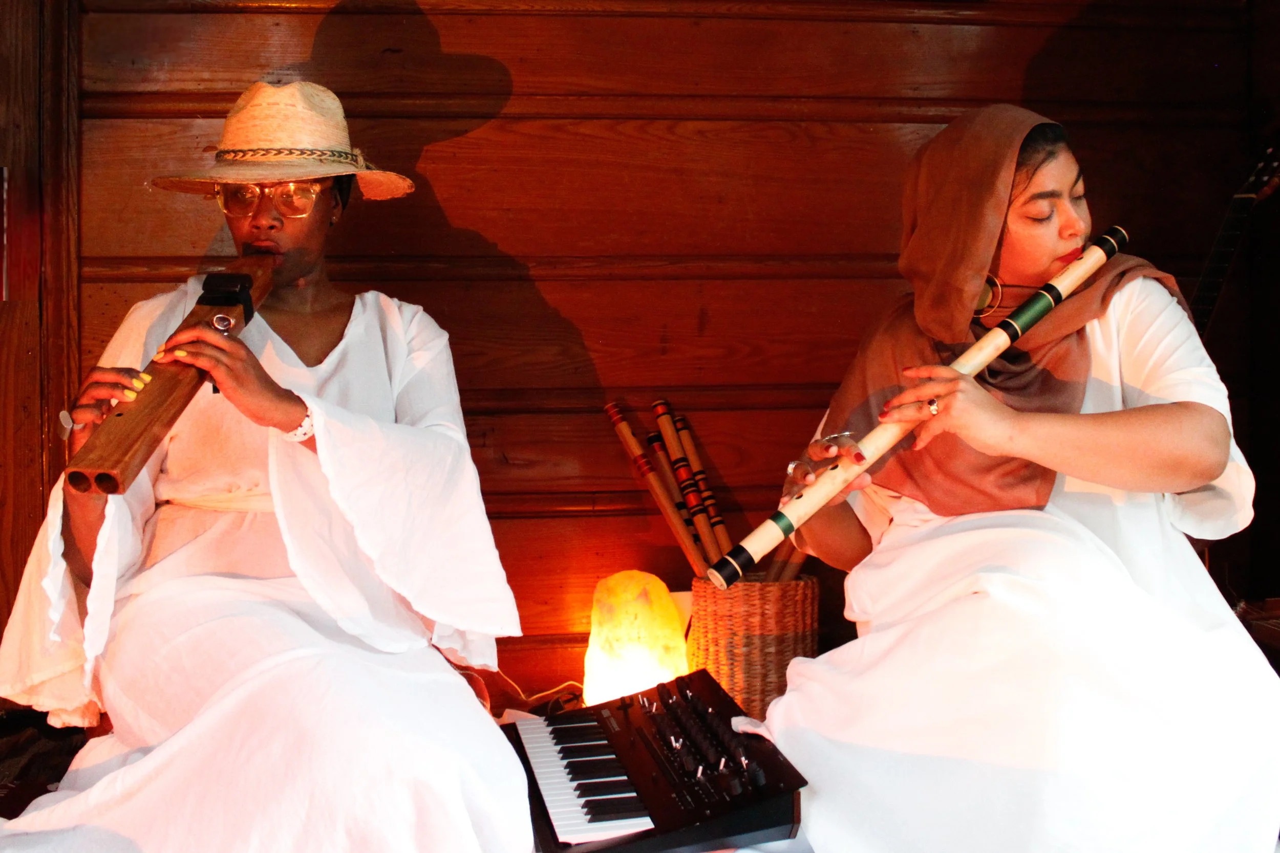 Two women sitting on the floor playing bamboo flutes, with a small keyboard and a salt lamp between them, against a wooden wall background.