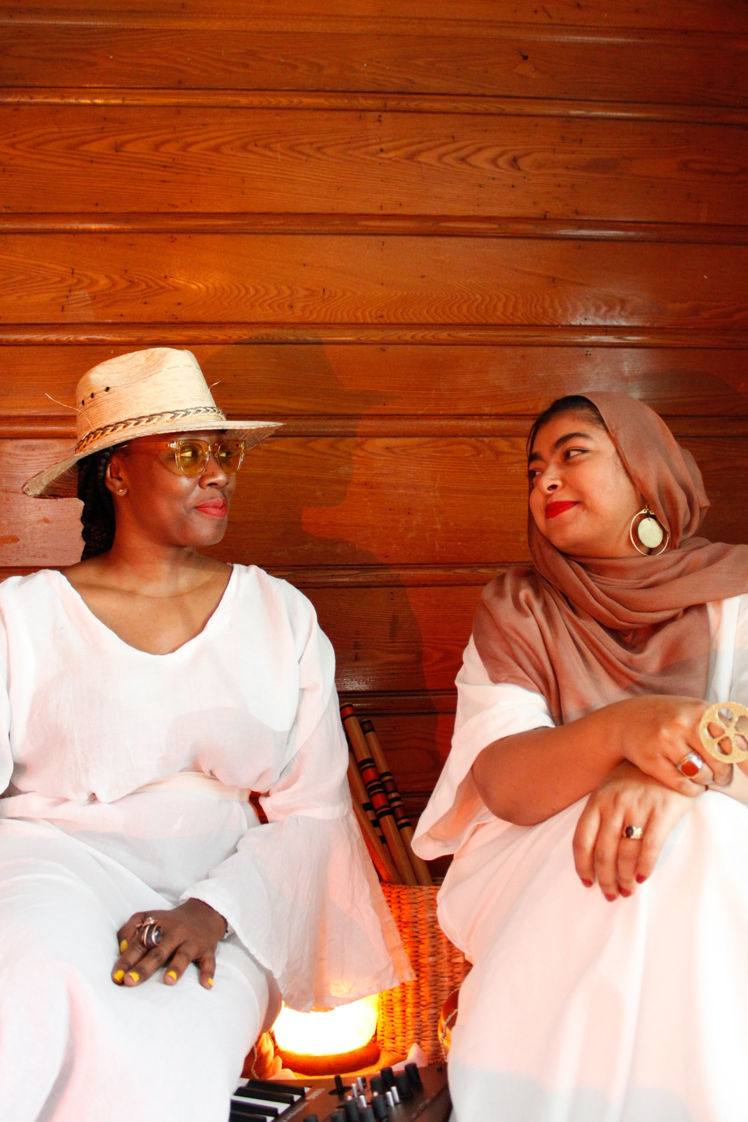 Two women sit and look at each other in front of a wooden wall, wearing white outfits and jewelry, with a small light and a keyboard on the floor between them.