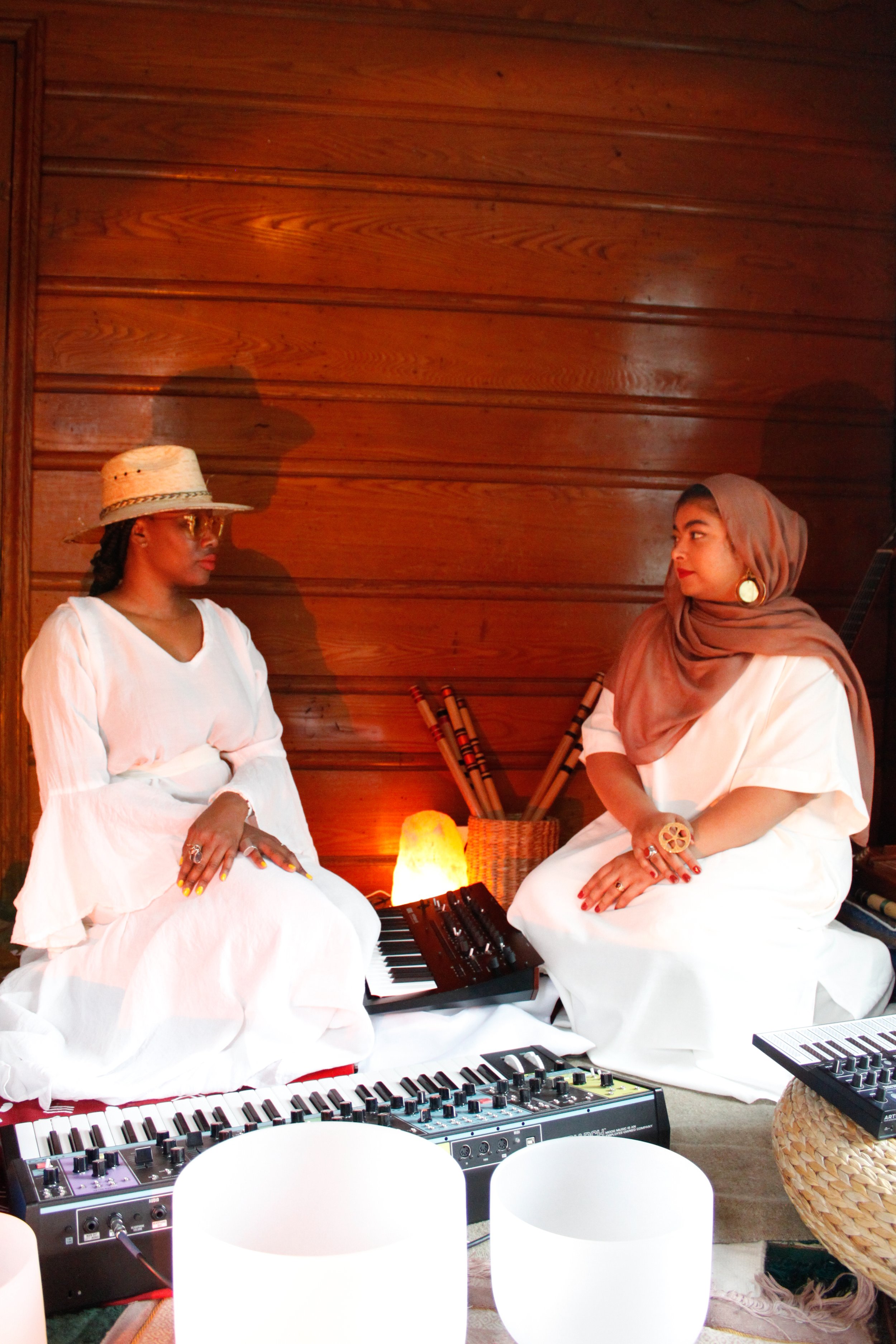 Two women dressed in white, sitting on the floor with musical equipment, in a room with wooden walls, engaged in a conversation.