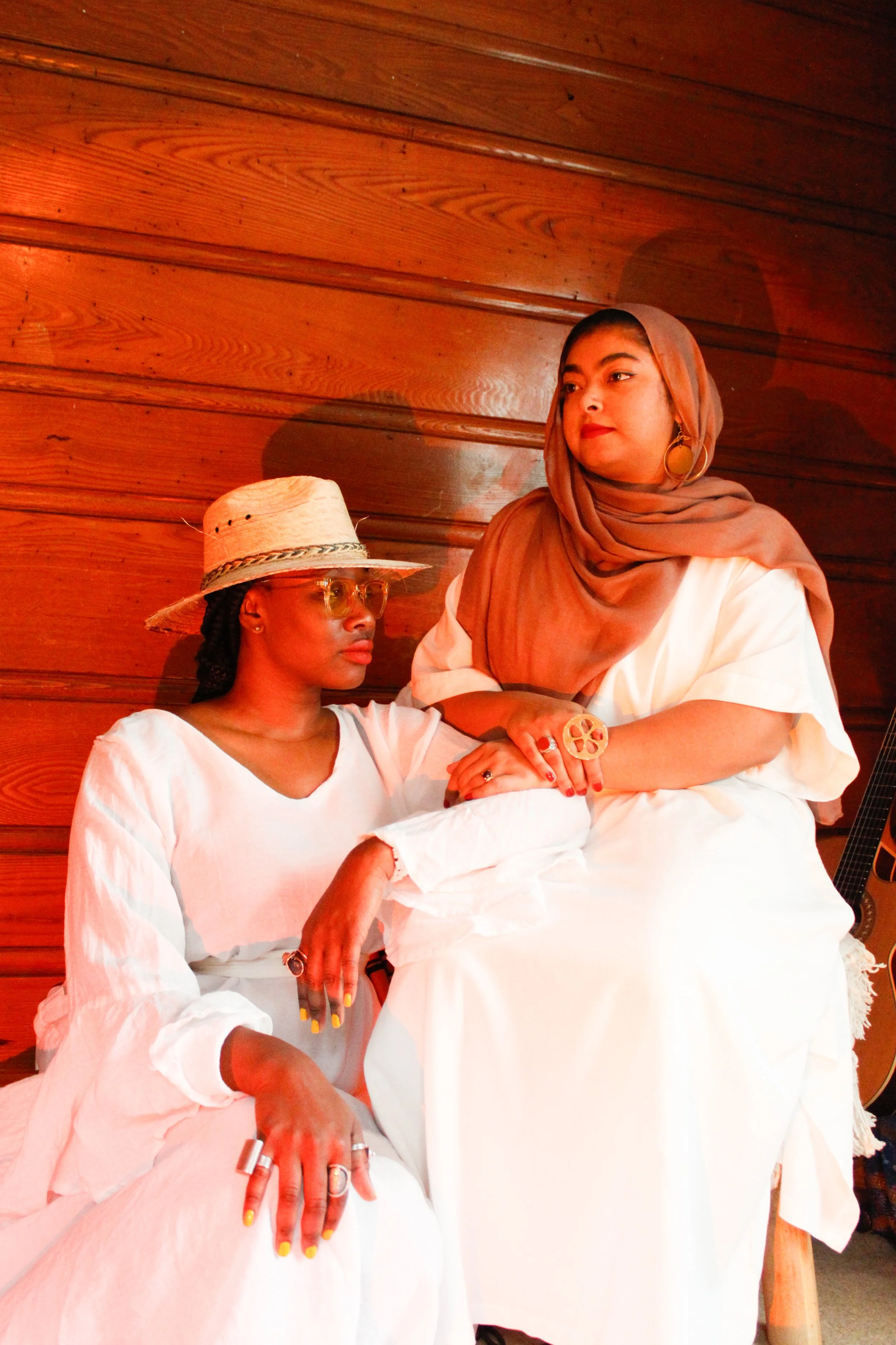 Two women in white dresses sitting against a wooden wall, one wearing a straw hat and glasses, the other with a brown headscarf and large jewelry.