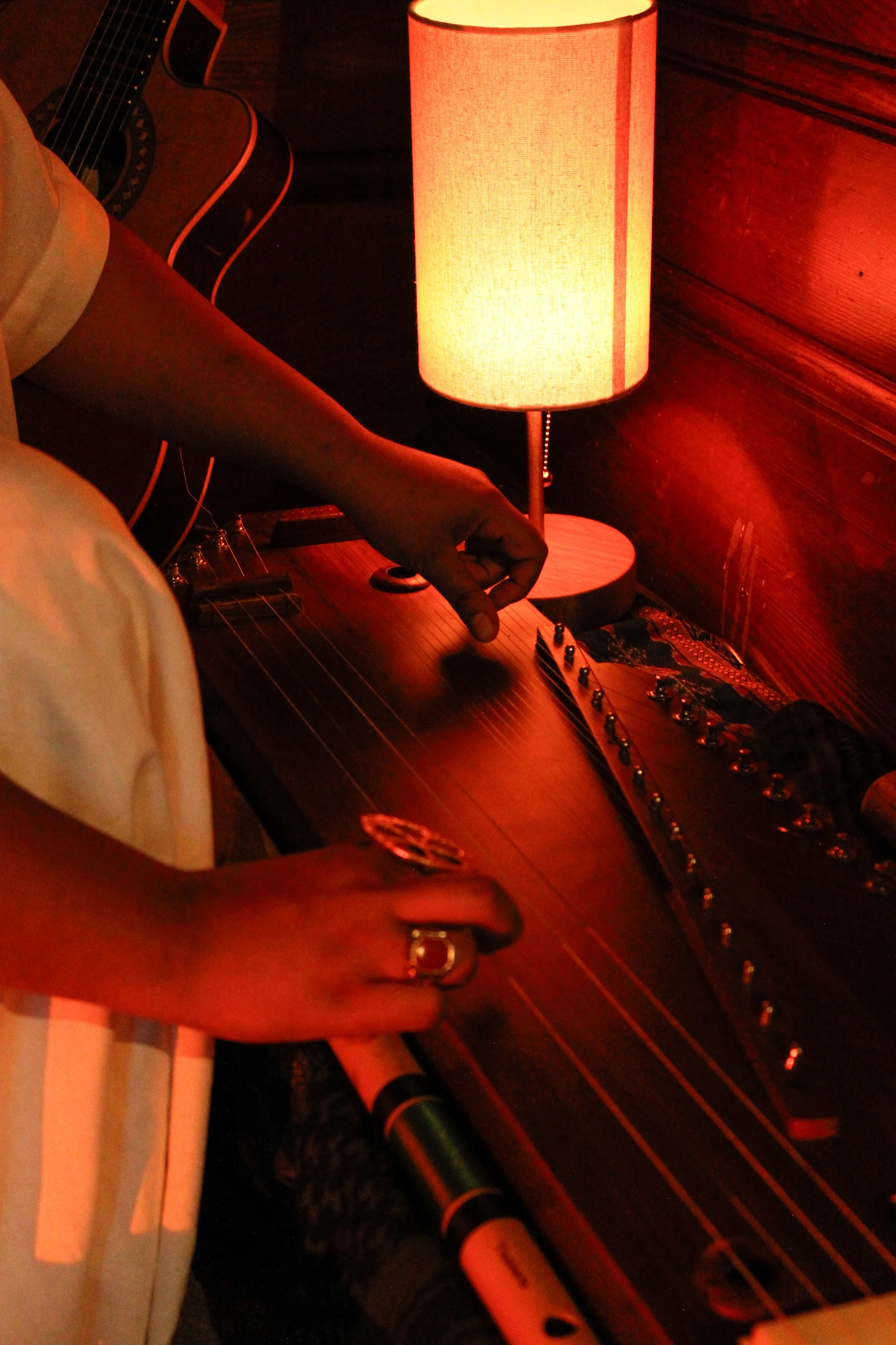 Person playing an acoustic guitar, with a lamp on a wooden surface nearby, in a dimly lit room.