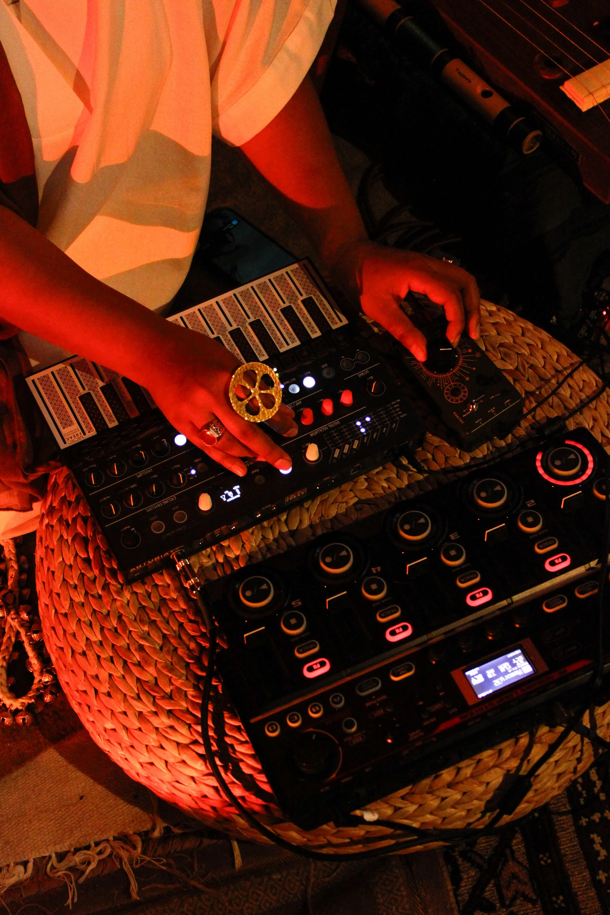 Person DJing with a mixer and controller, wearing a large gold ring, on a woven stool with red lighting
