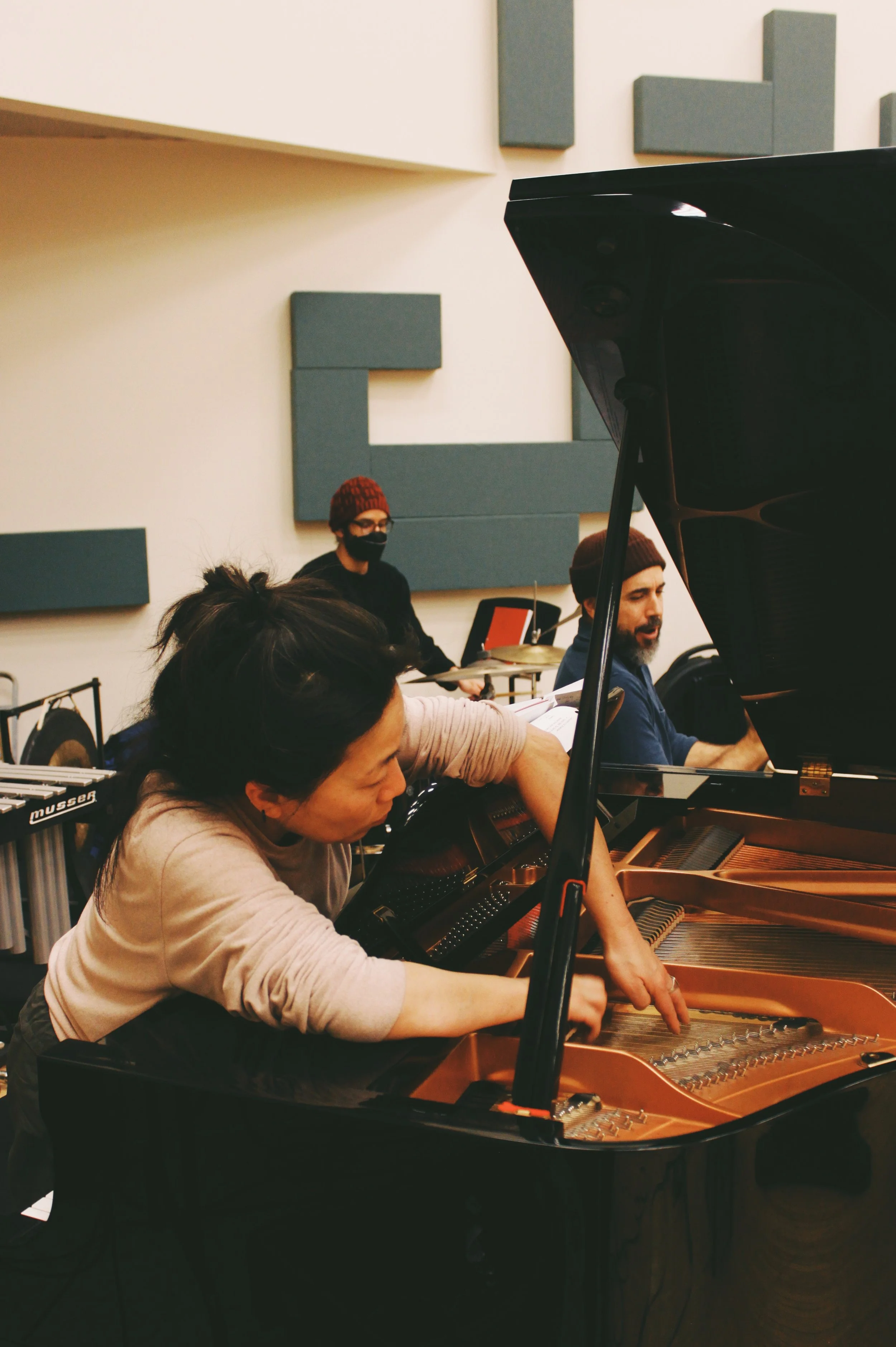 Three musicians, two men and one woman, rehearsing at a grand piano, with a percussionist playing drums in the background.