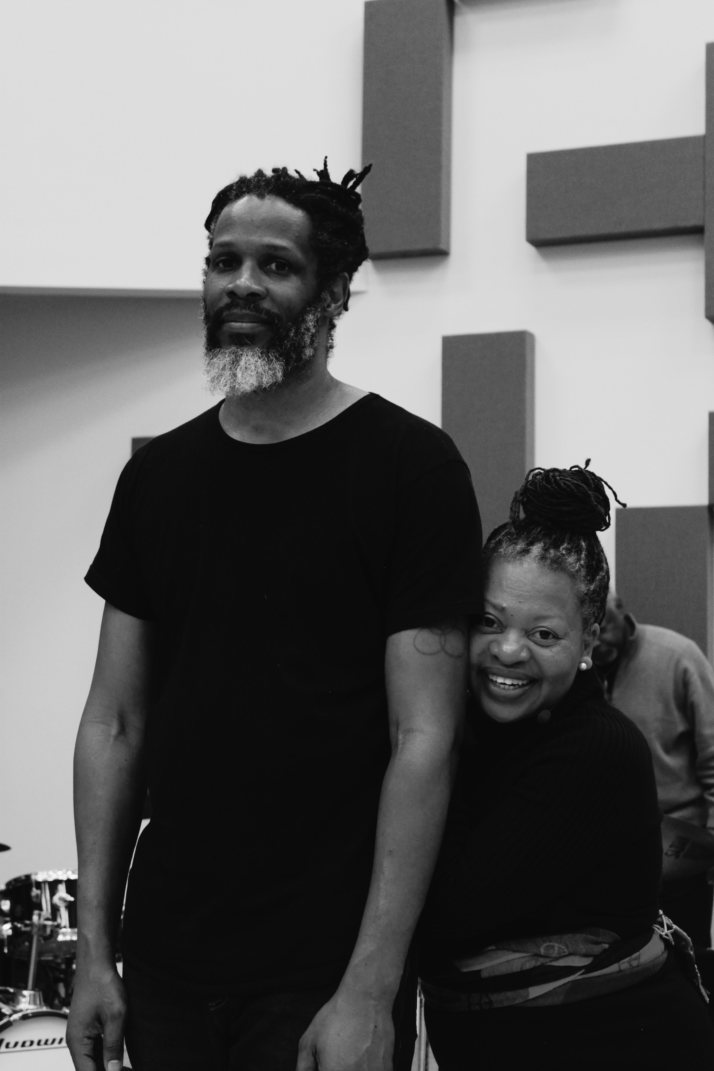 A black-and-white photo of a man with locks and a beard standing next to a woman with locks in a bun, smiling and hugging his arm. They are in a room with soundproofing foam panels on the wall.