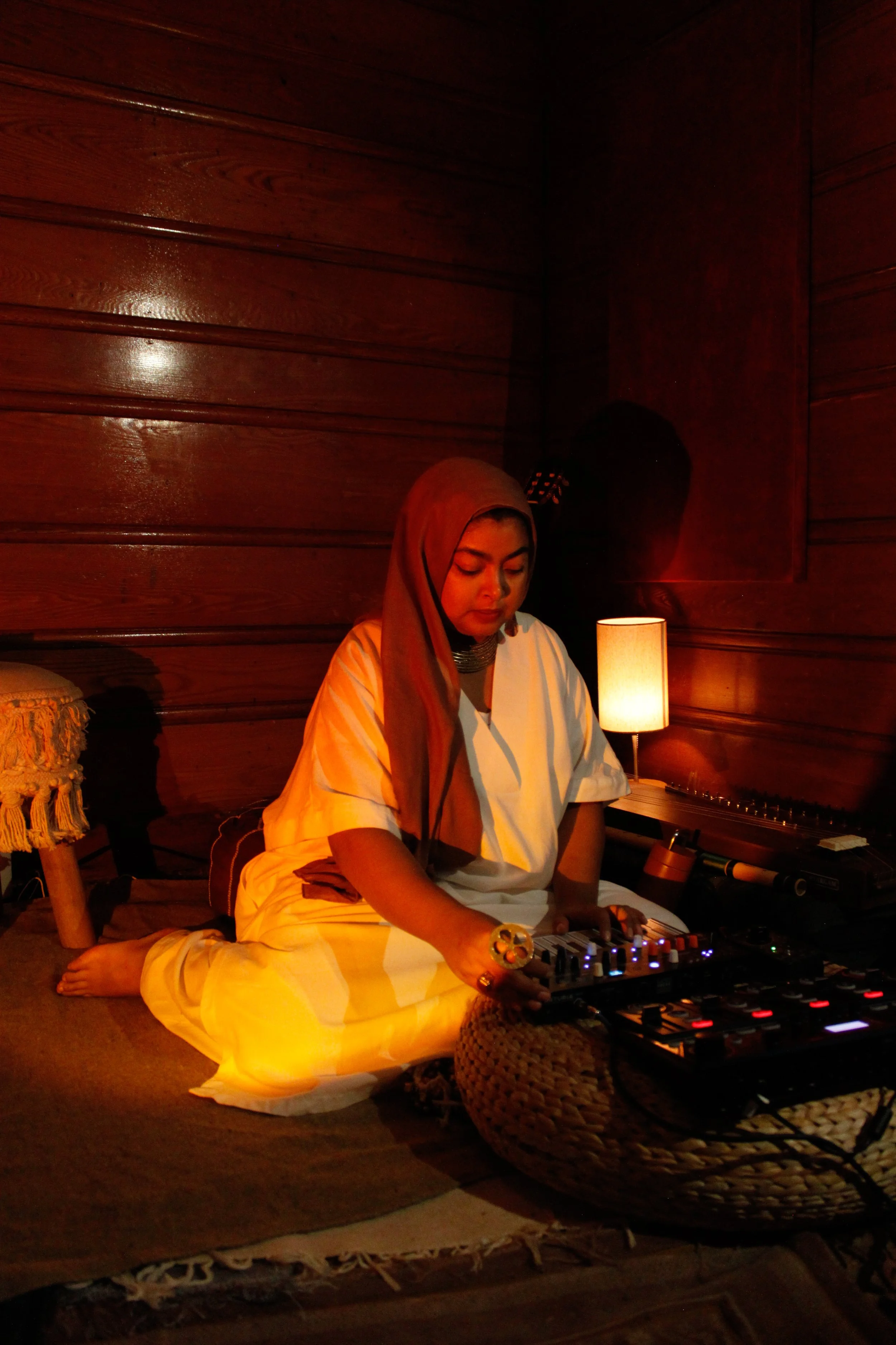 A woman sitting on a mat in a dimly lit room with wooden walls, surrounded by electronic music equipment and a small table lamp.