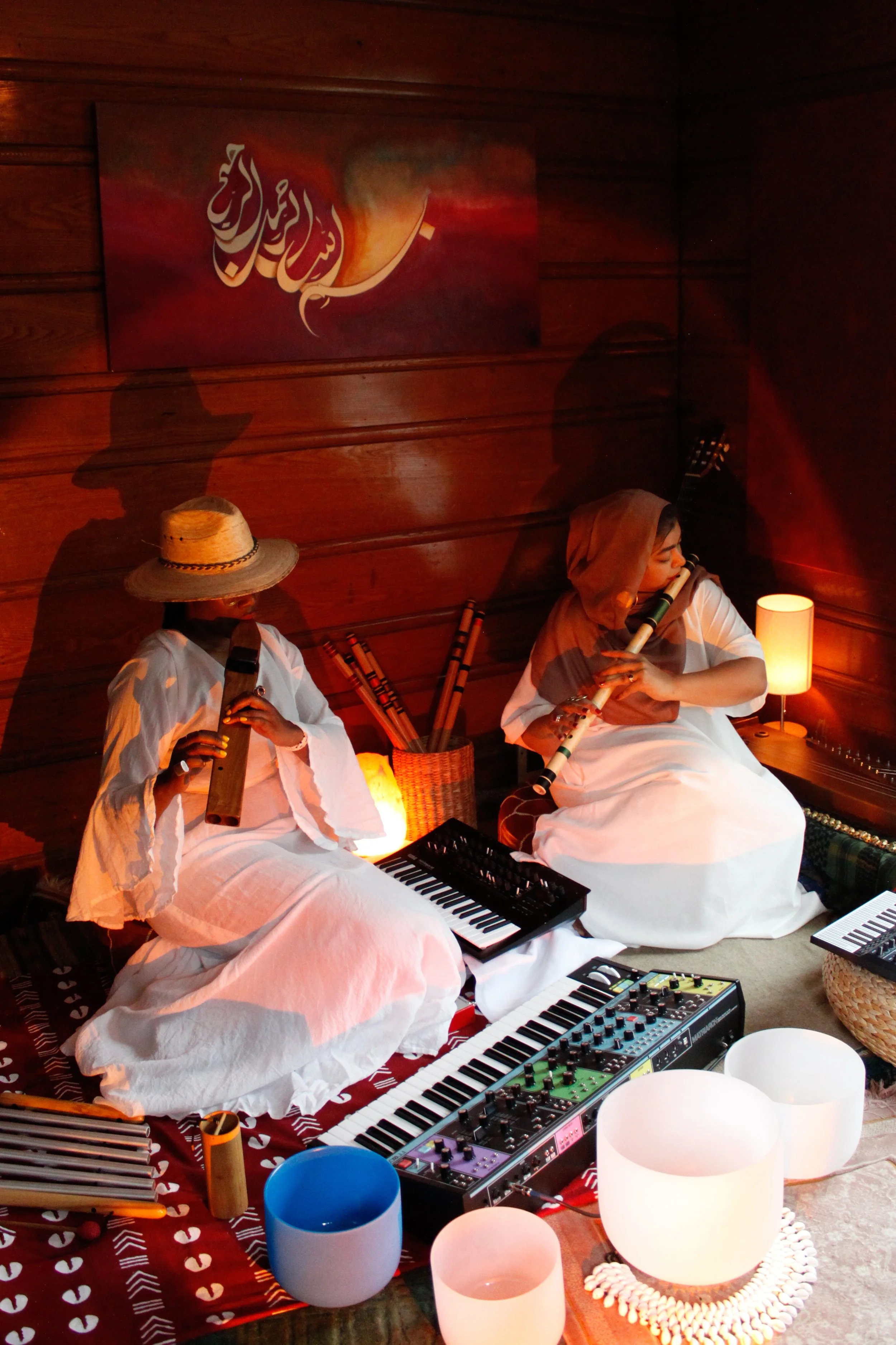 Two women in white dresses playing musical instruments, one with a hat and the other with a headscarf, seated on a carpeted floor with musical equipment and instruments, illuminated by warm lighting in a cozy wooden room.