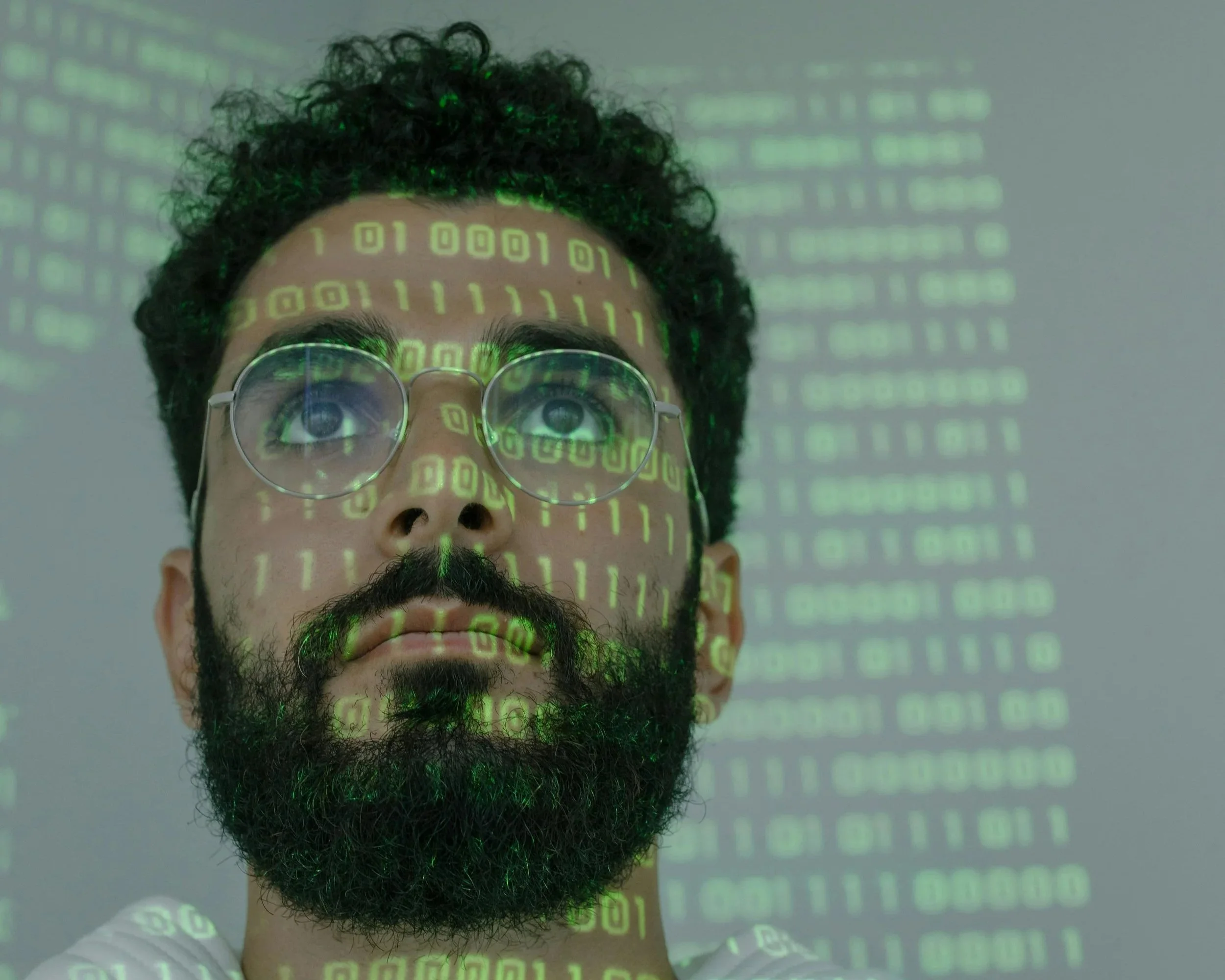 A man with glasses and a beard looking up. Green binary code is projected onto his face and the background.