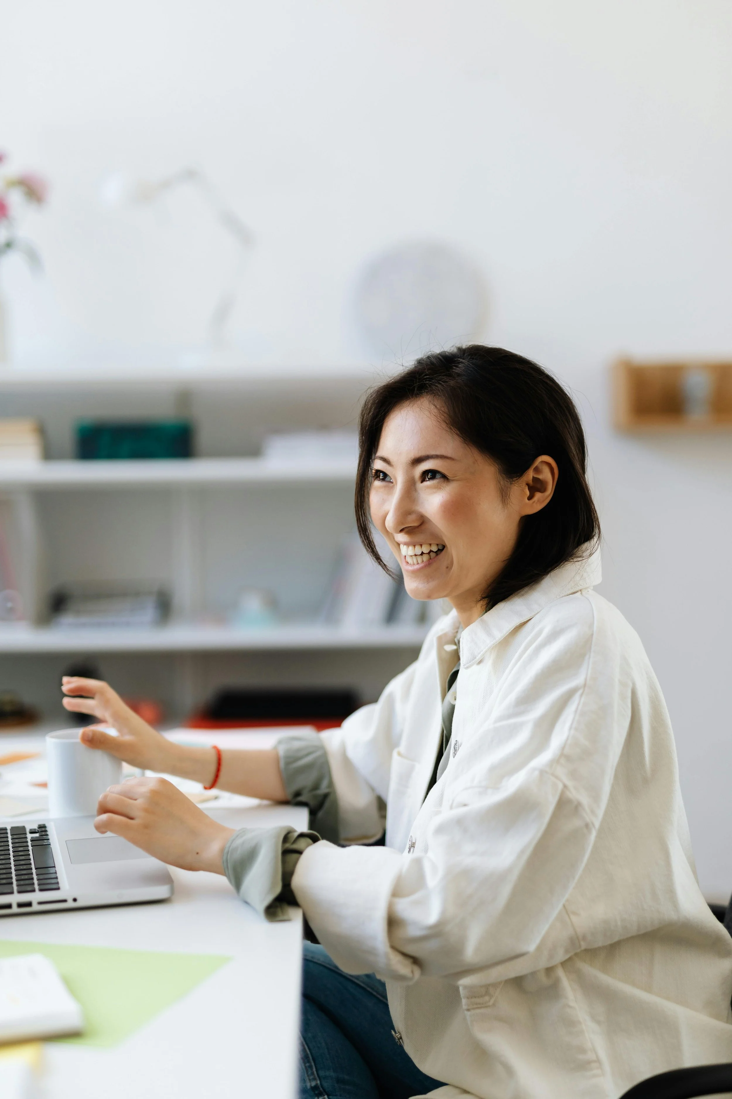 A woman with shoulder-length dark hair, smiling and gesturing with her hand, sitting at a desk with a laptop and a mug in a bright, modern office.