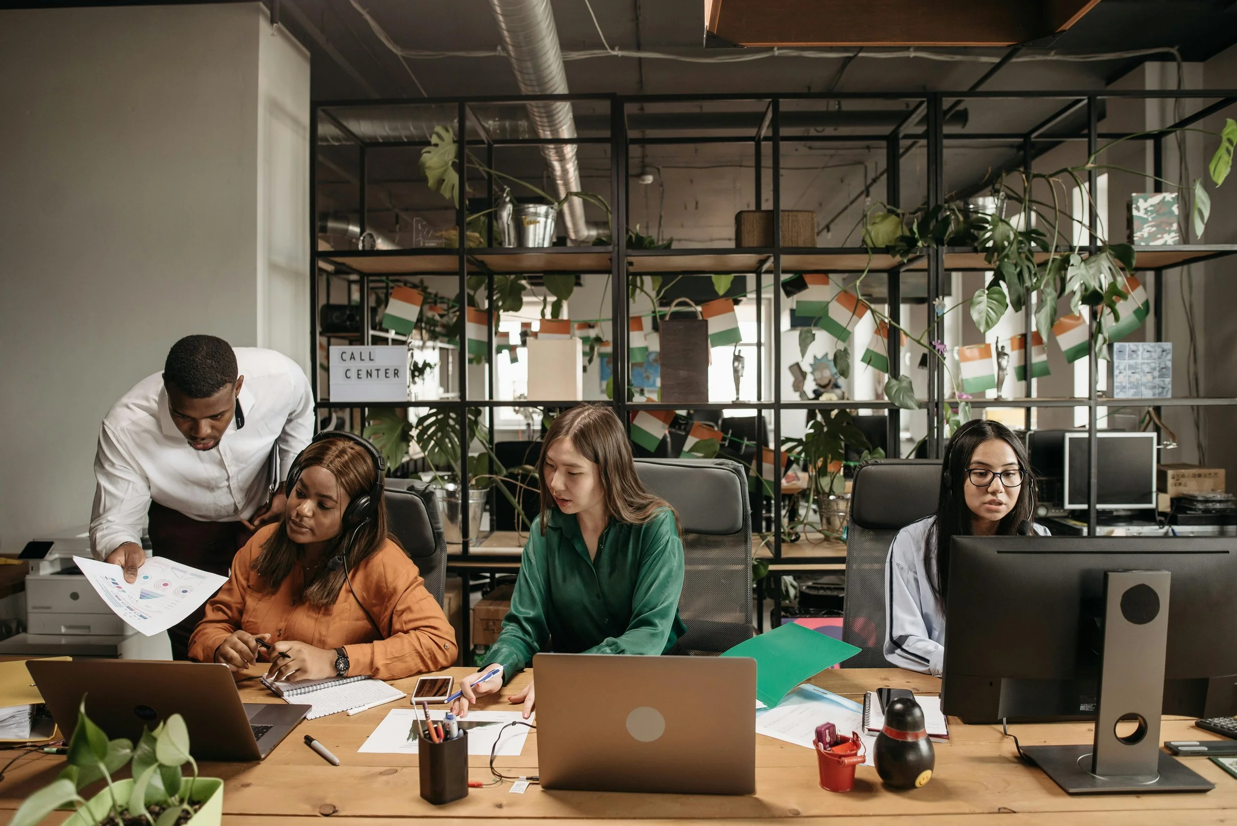 A busy office with four women working at their desks, with one man standing and talking to a seated woman, in front of a shelving unit decorated with plants, charts, and a sign that says 'Call Center'.