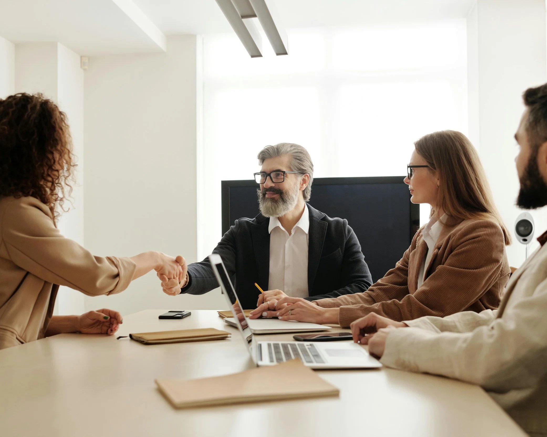 Business meeting with two people shaking hands across a conference table, surrounded by three others with laptops and notebooks, in a bright office.