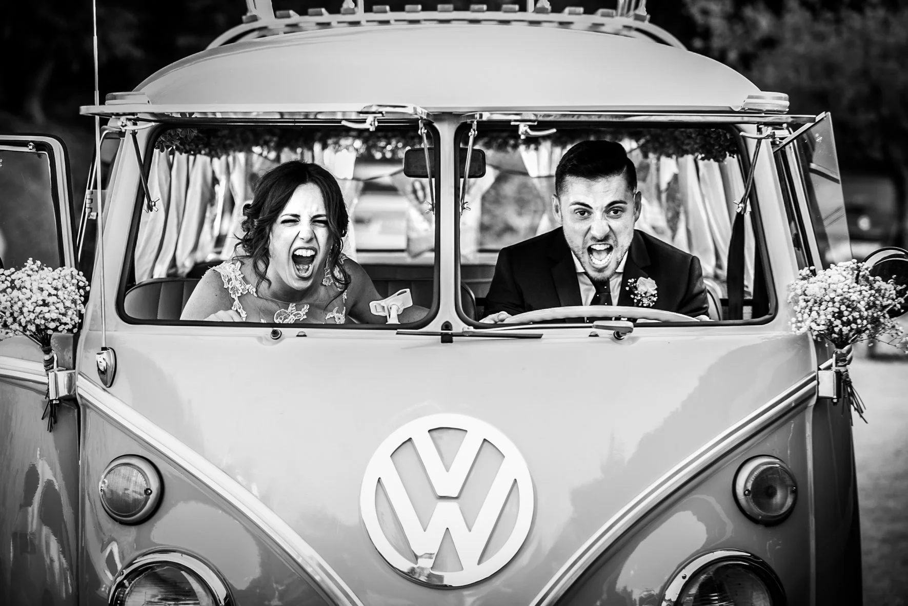 A black-and-white photo of a woman and a man inside a vintage Volkswagen van. The woman has dark hair styled up and appears to be yelling or singing with a wide-open mouth, wearing a lace dress. The man has short hair, is dressed in a suit, and is also yelling or singing with a wide-open mouth, holding the steering wheel. The van is decorated with flowers on the sides, suggesting a festive or celebratory occasion.
