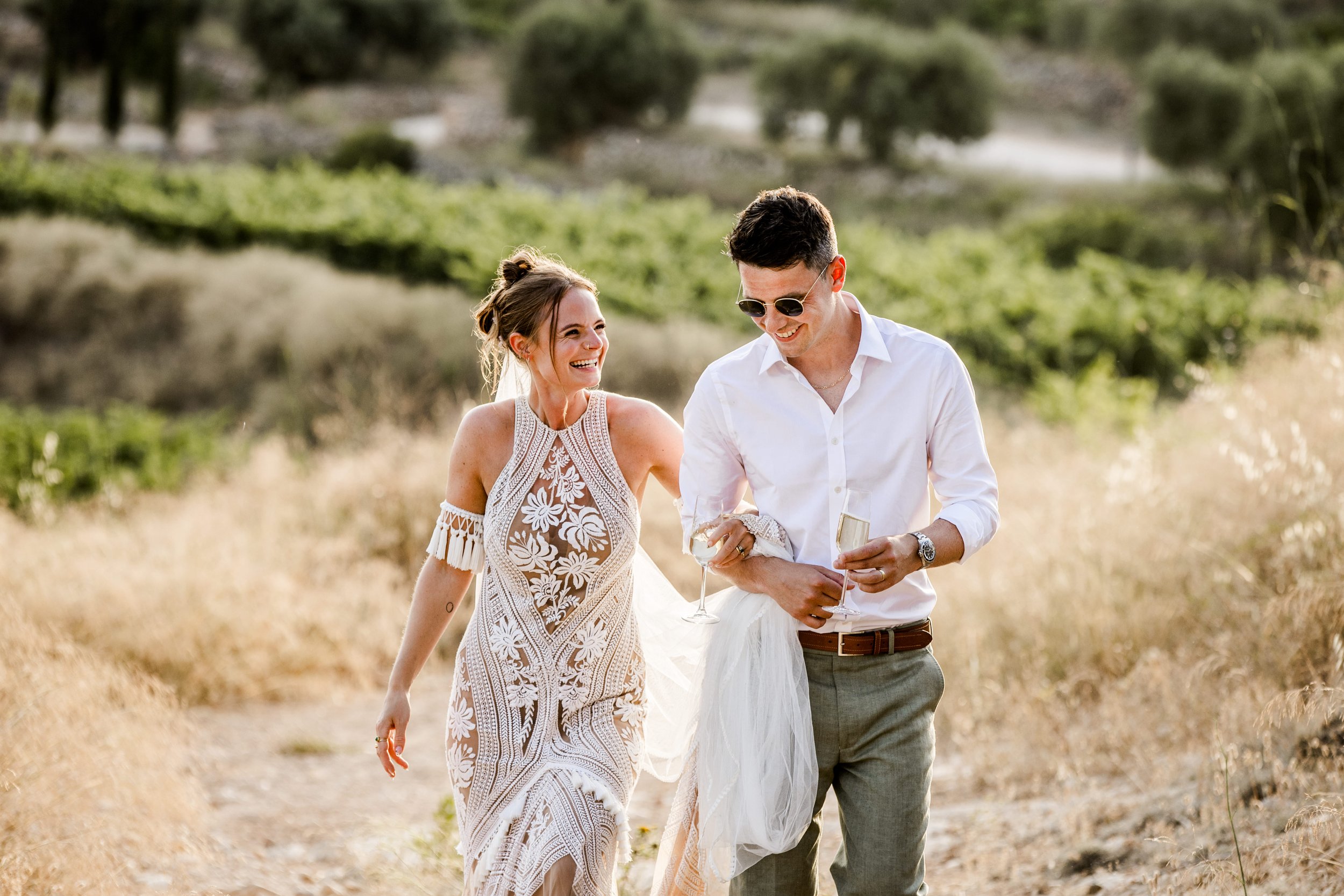 A smiling woman and man walking outdoors in a sunny field, holding glasses of wine. They look happy and are enjoying a relaxed moment together during their destination wedding in Masia Cabellut, Spain