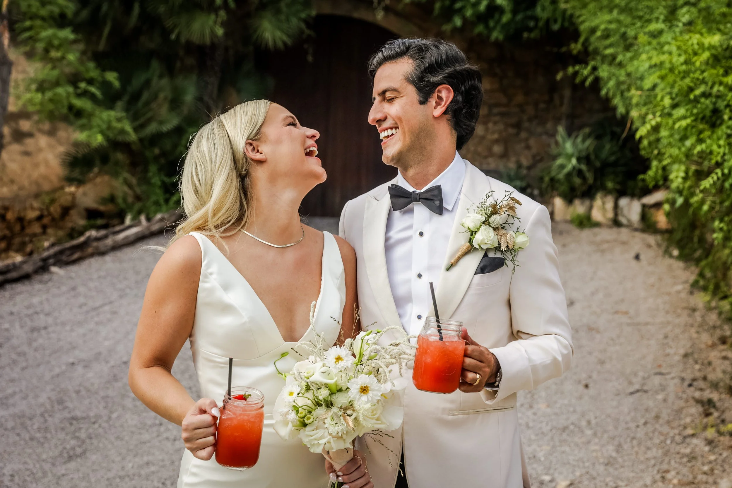 A smiling bride and groom in wedding attire sharing a joyful moment outdoors, holding drinks in mason jars and a bouquet of white flowers.