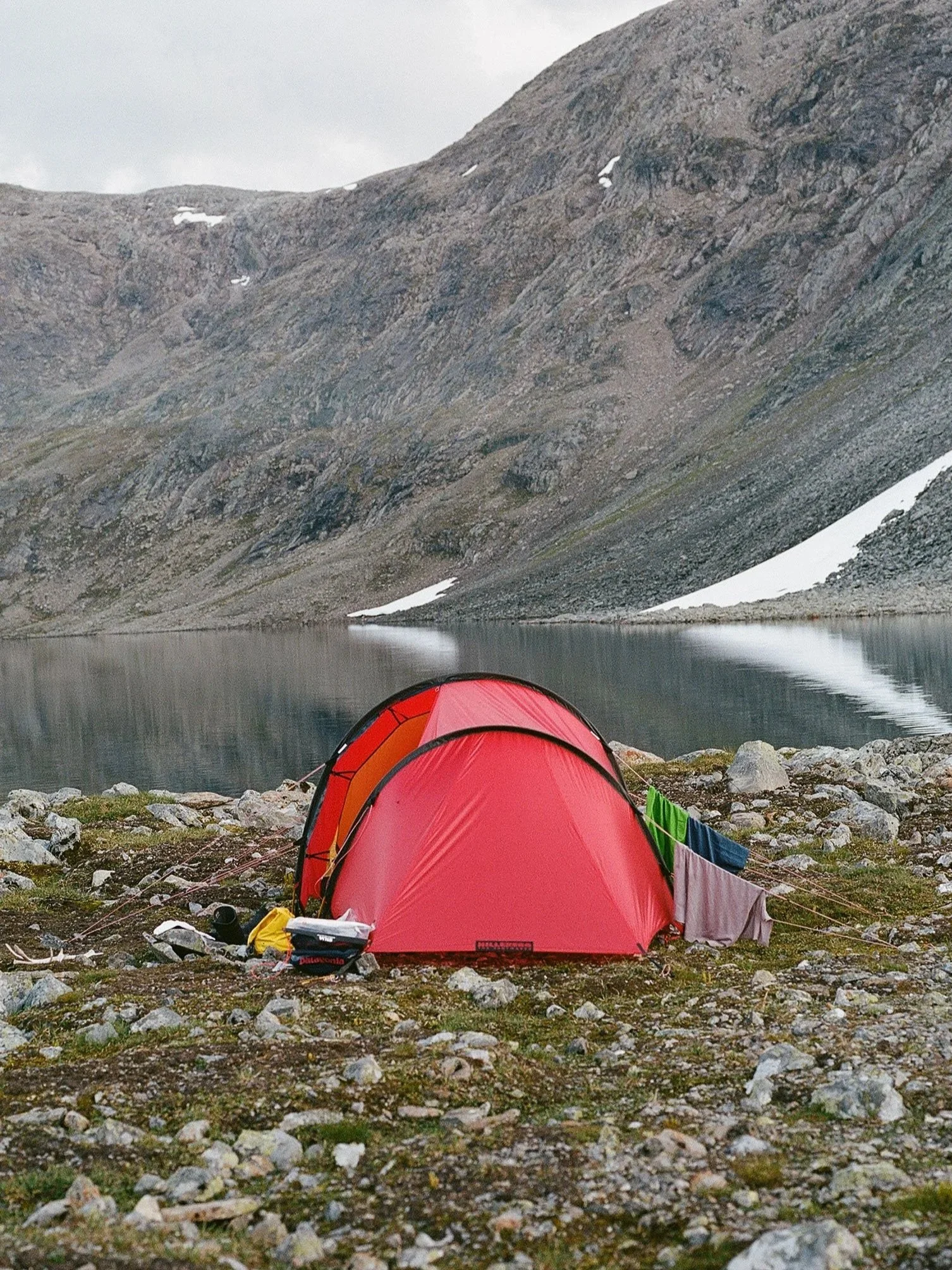 A red camping tent set up on rocky ground beside a calm mountain lake with mountainous terrain in the background, and some clothing hanging on a line attached to the tent.