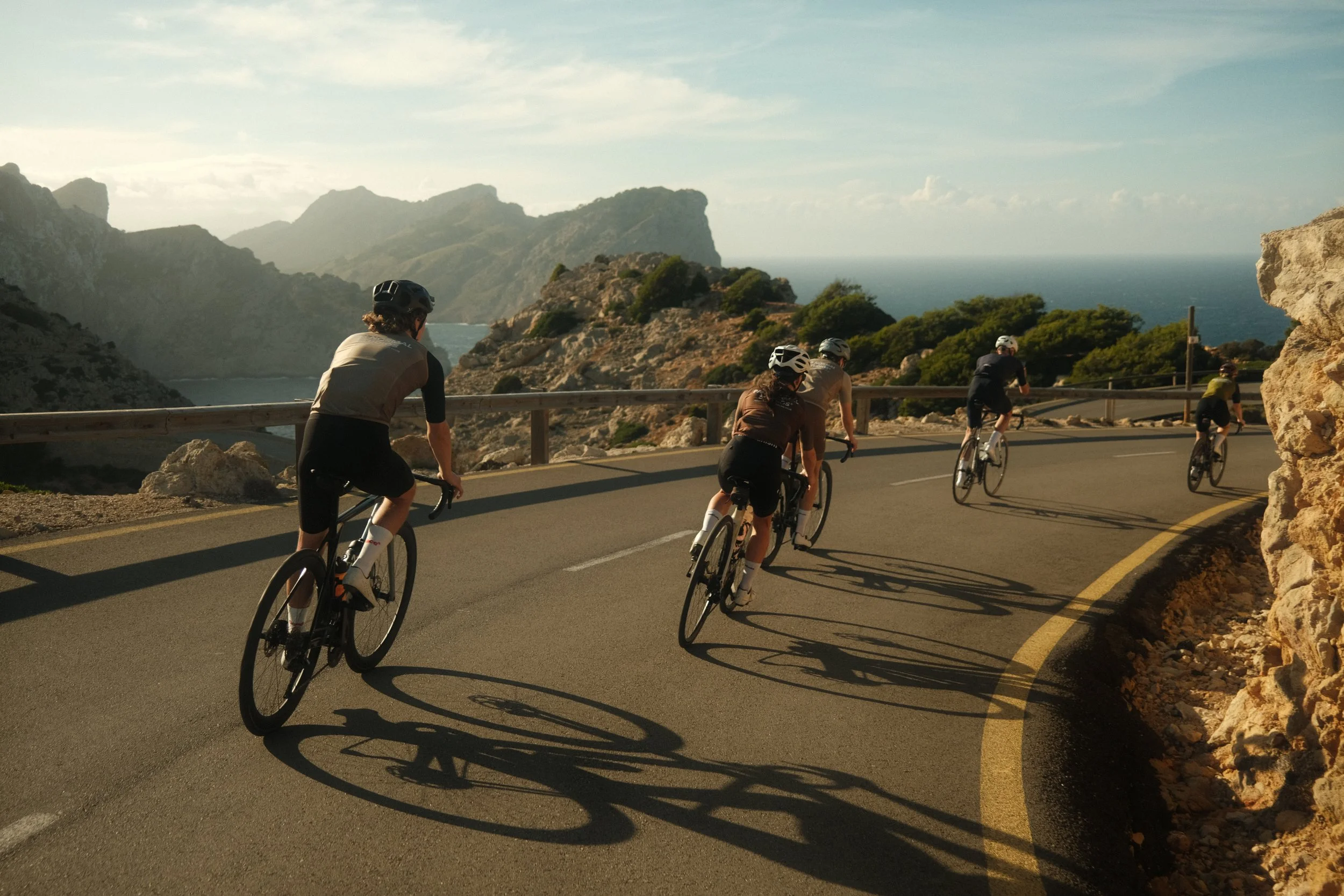 Group of five people riding bicycles on a winding coastal road with mountains and ocean in the background during daytime.