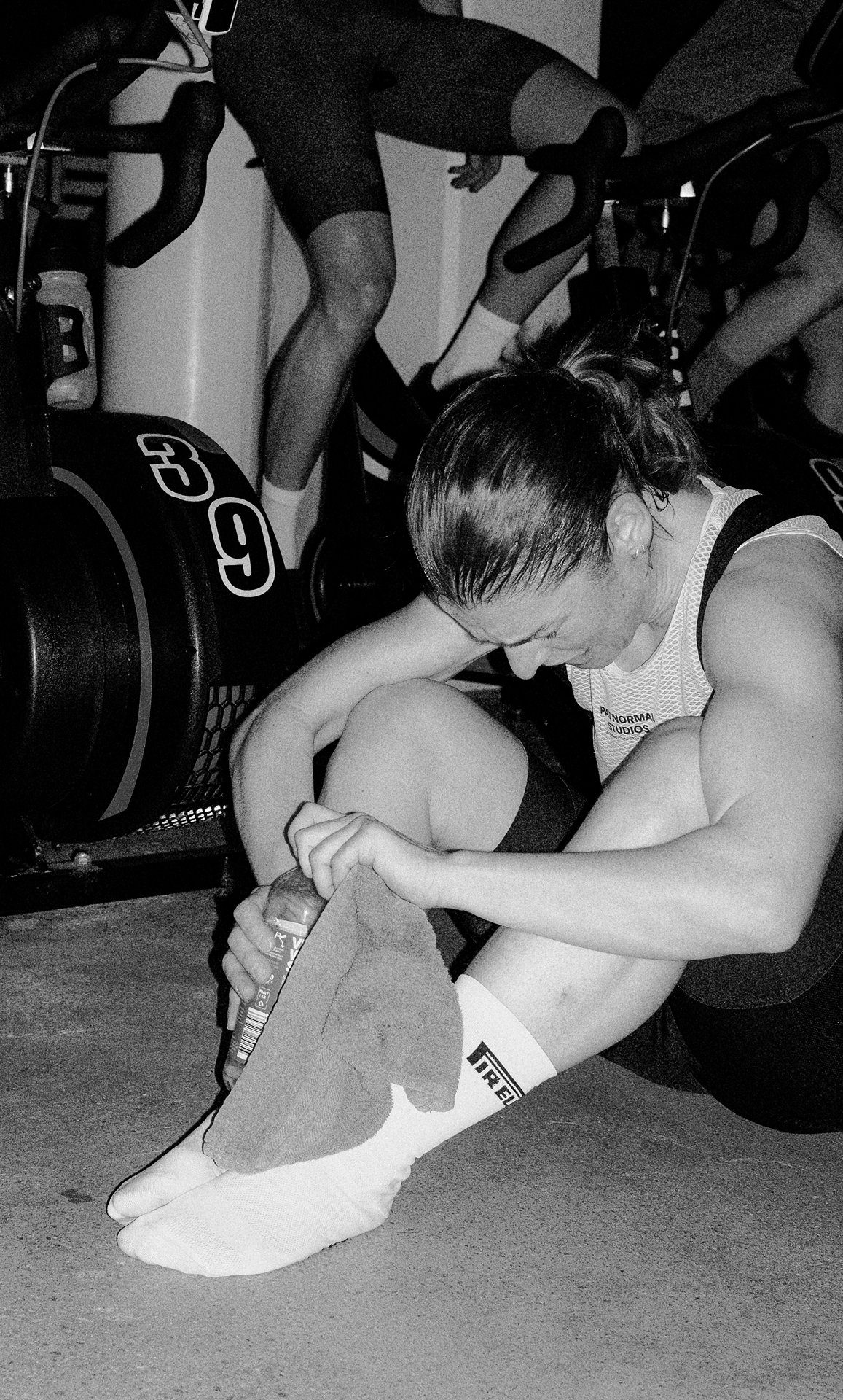 A woman sitting on the floor in athletic attire, holding a towel and water bottle, appears exhausted after a workout, with exercise bikes and other people in the background.