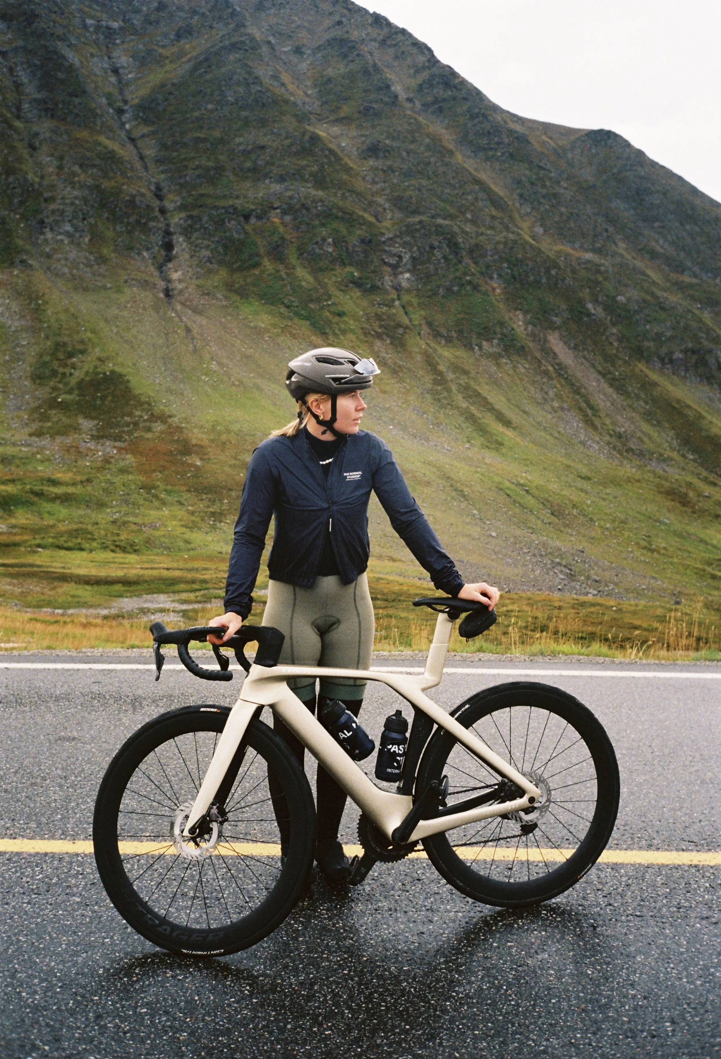 A woman with a helmet standing next to her beige road bicycle on a wet road, with rolling green hills and mountains in the background.