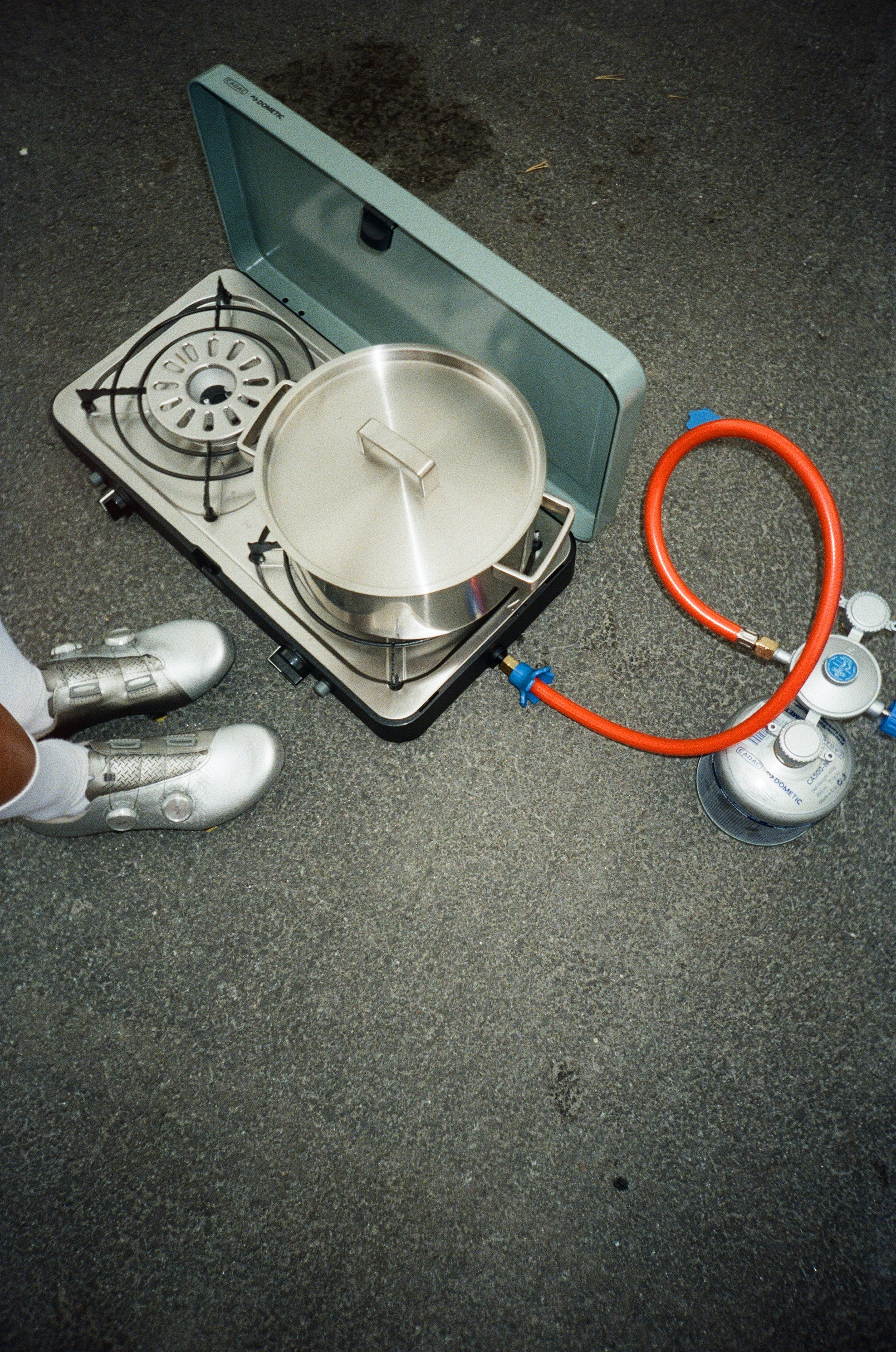 A portable gas stove with a large cooking pot on it, connected to a gas cylinder with a red hose. A person wearing white sneakers is standing nearby on a rough concrete surface.