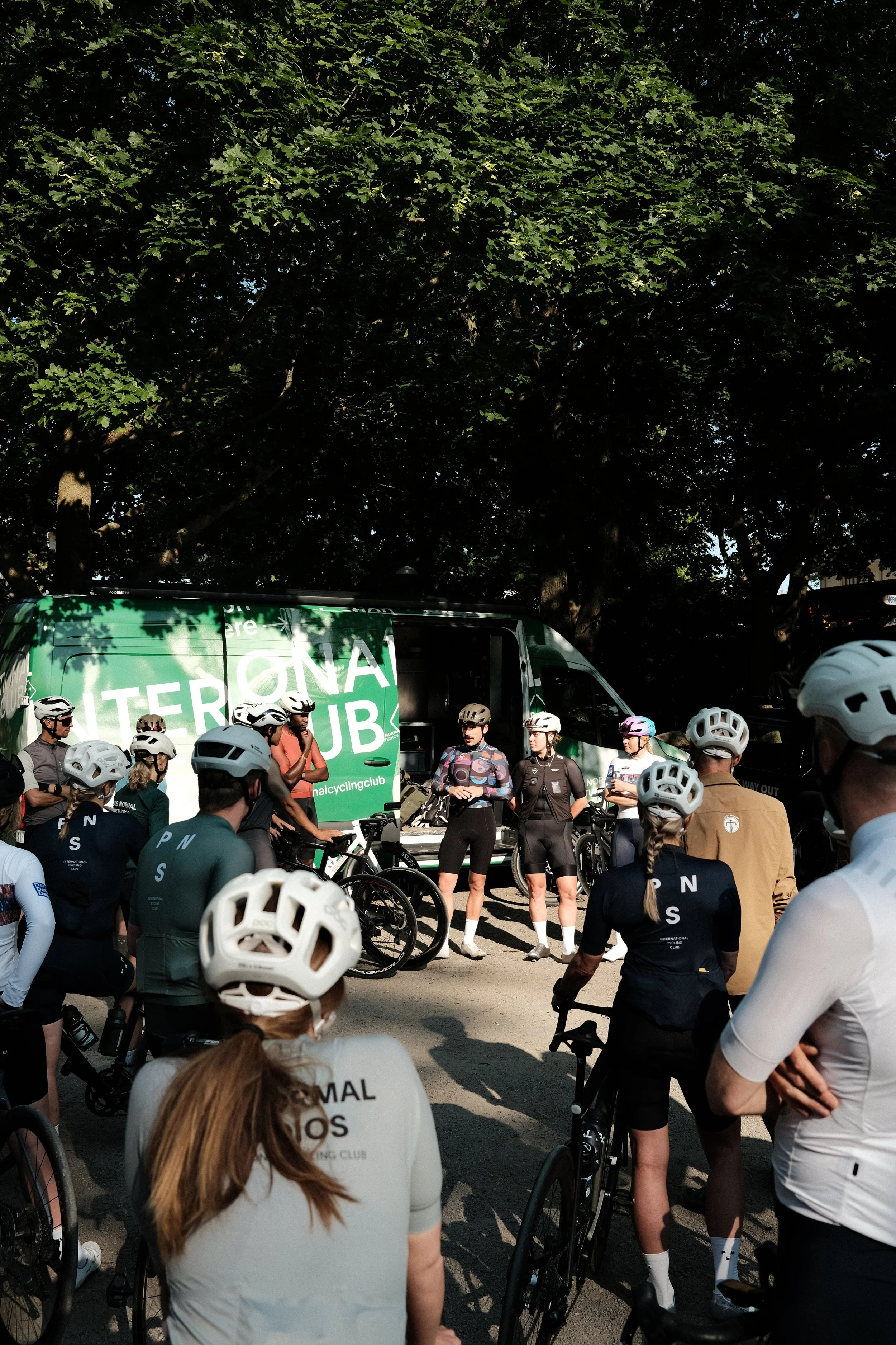 Group of cyclists gathered outdoors, wearing helmets and sports gear, with a green vehicle in the background displaying 'INTERNATIONAL CYCLING CLUB'.