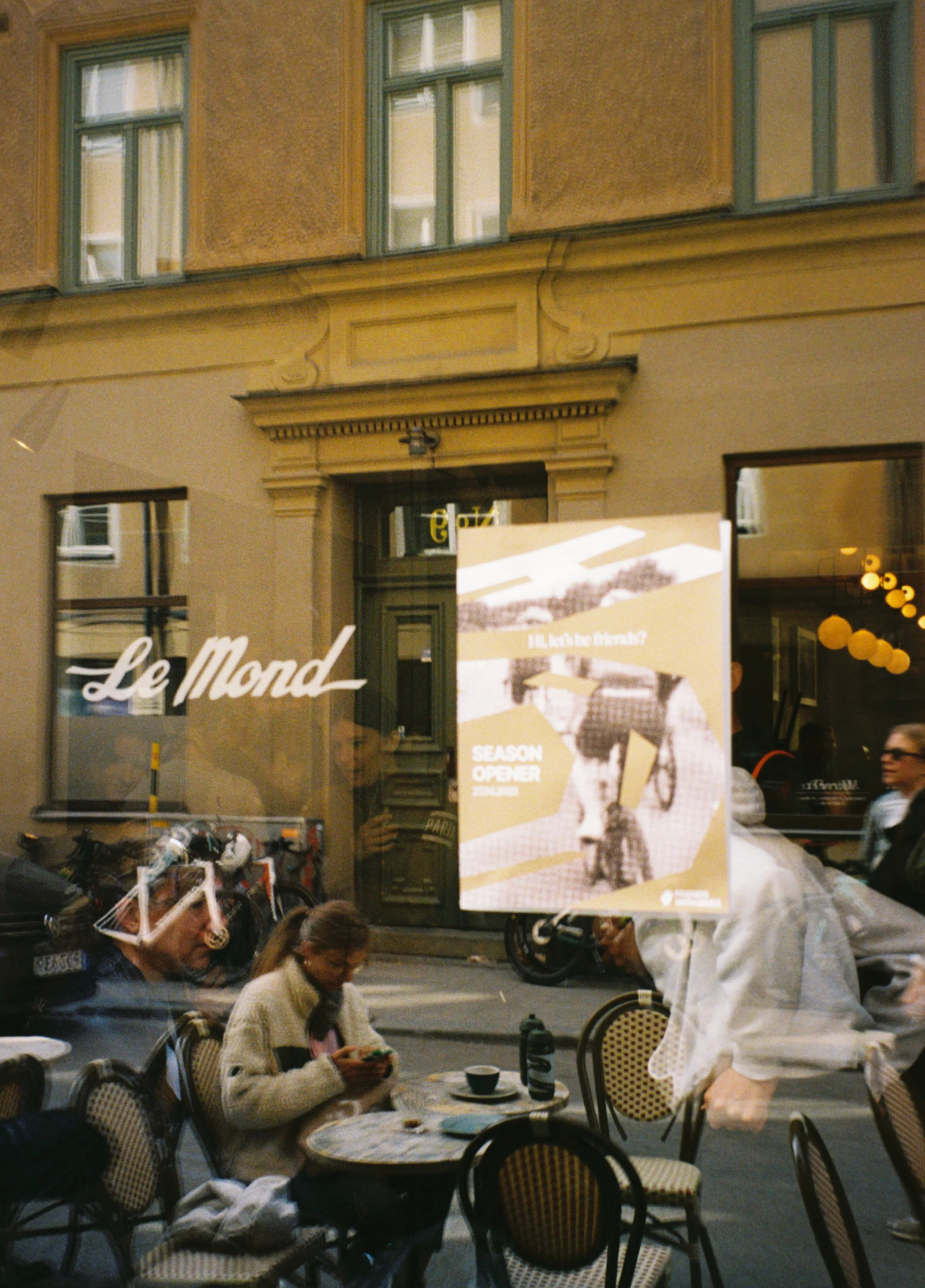 Street scene viewed through a window reflecting an outdoor café with people seated at tables. A woman is looking at her phone while others are engaged in conversation. There is a sign for 'Le Mond' and a poster for a season opener event.