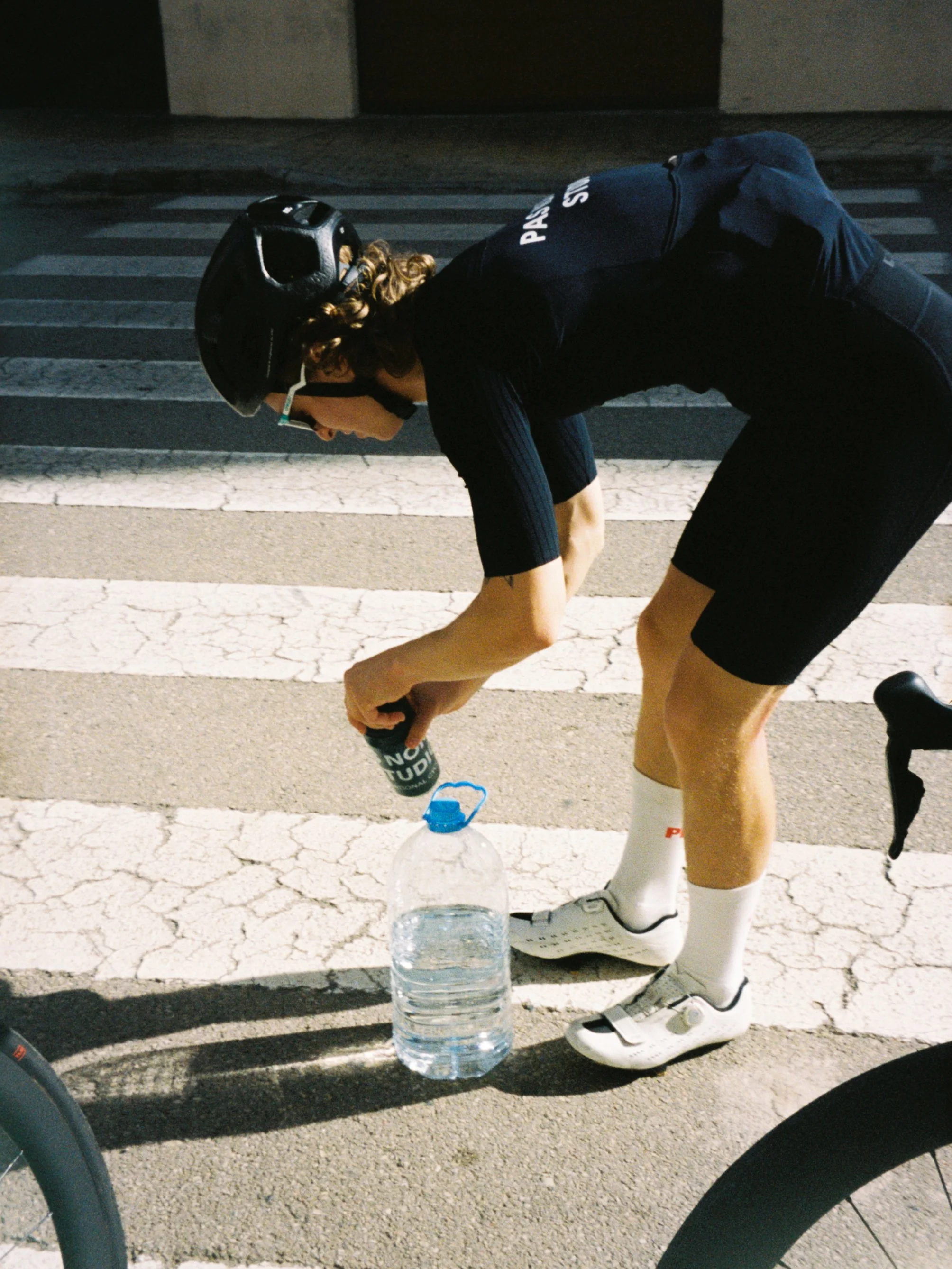A cyclist wearing a black helmet, black cycling jersey, black cycling shorts, and white cycling shoes, bending down on a crosswalk while pouring water from a bottle into a large plastic water container.