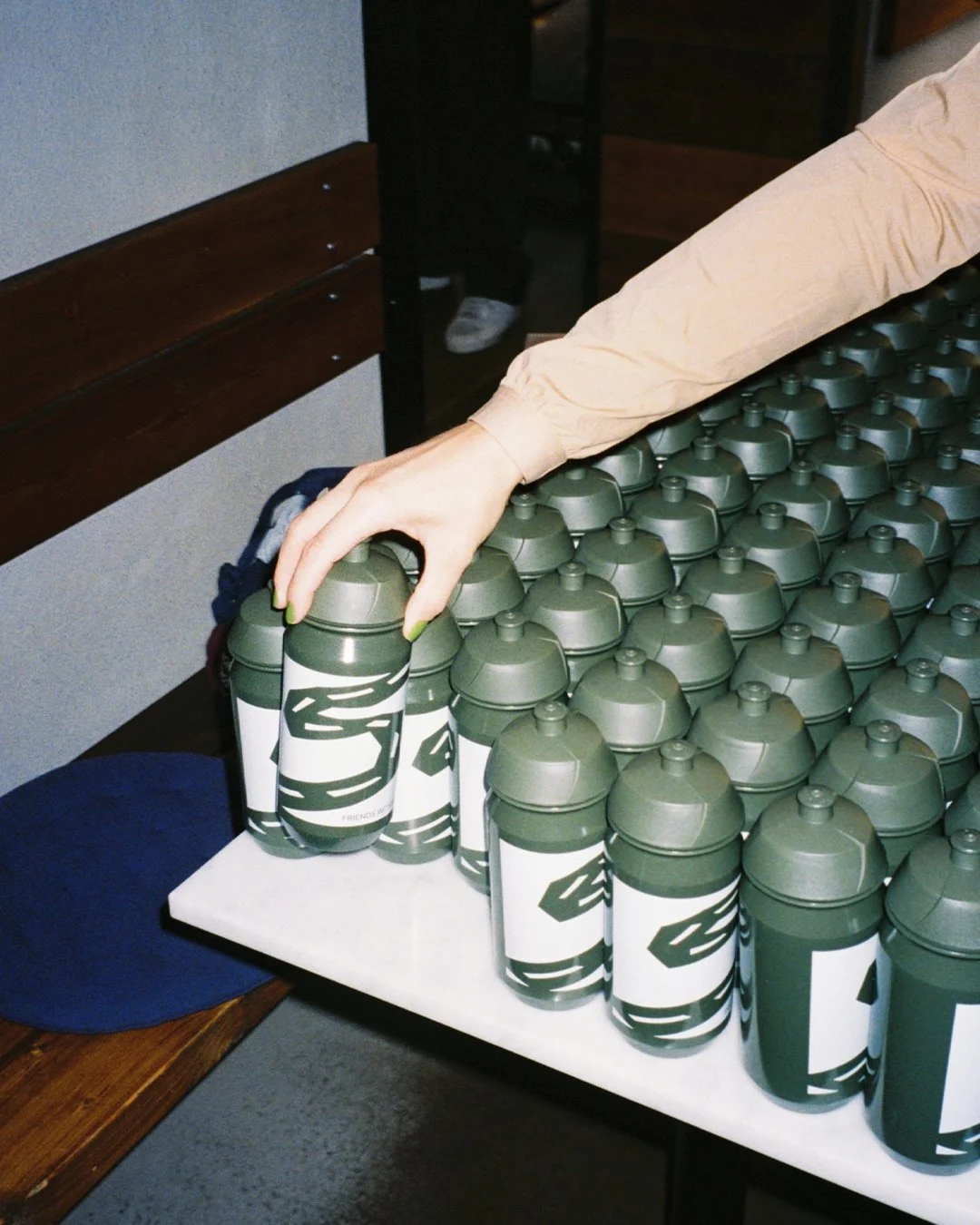 A person with pale skin, wearing a beige long-sleeve shirt, reaches to grab a green and white water bottle from a large display of similar bottles on a table.