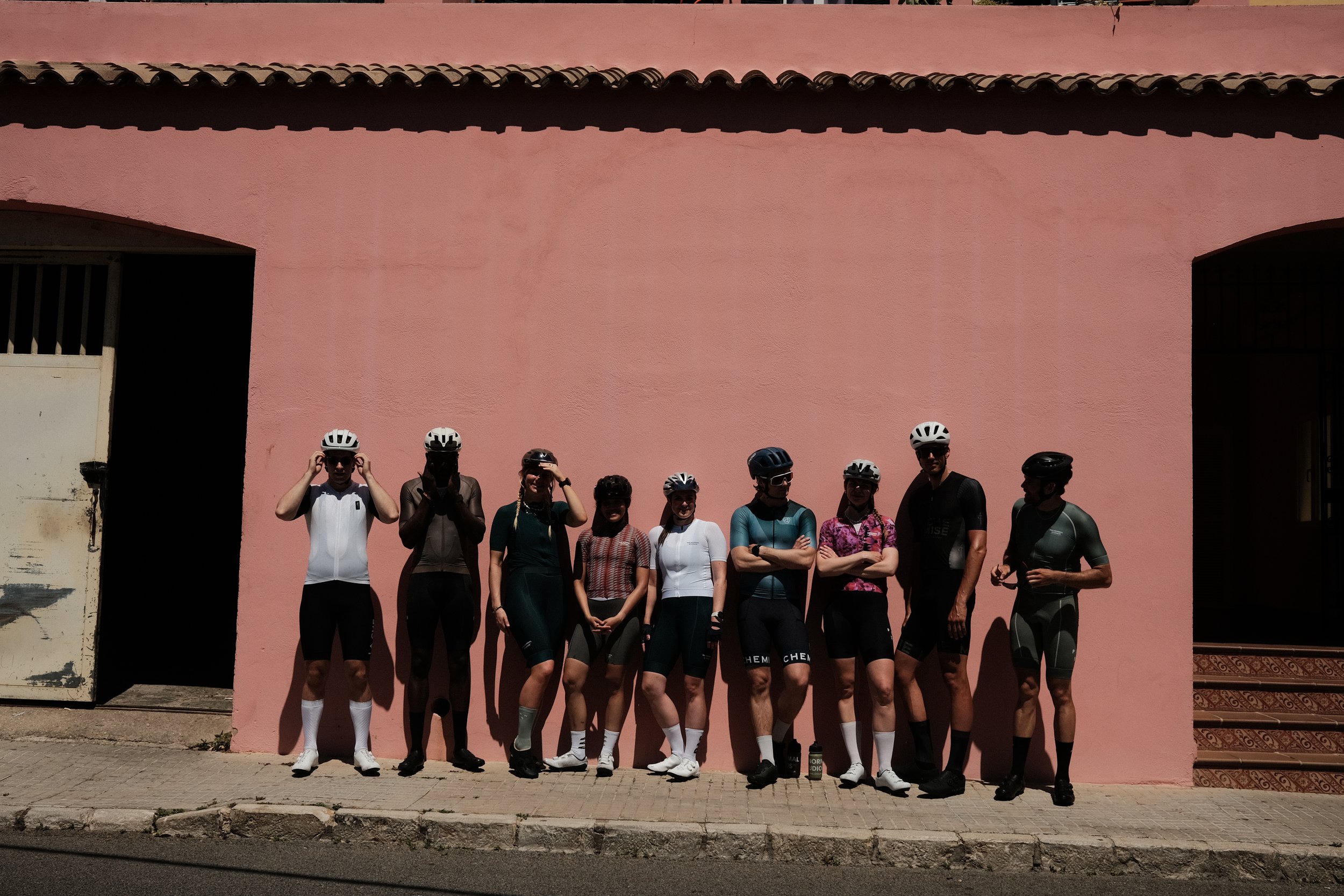 Group of ten cyclists standing against a pink wall, wearing helmets and cycling gear, some adjusting their helmets, with stairs and a door on the right.