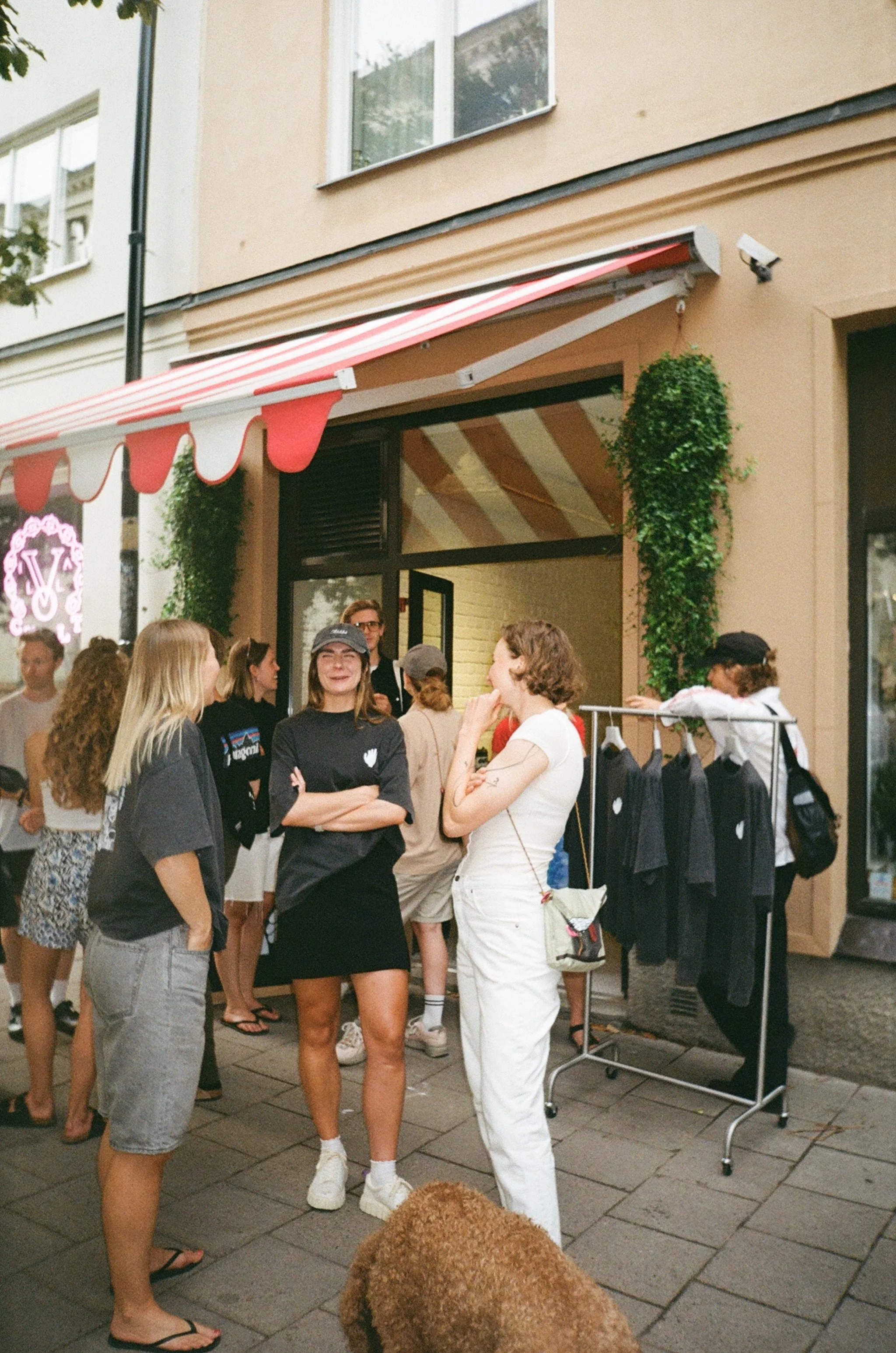 People standing and talking outside a storefront with a red and white striped awning; clothing items hanging on a rack; a person with a dog in front; neon pink letter sign visible on the storefront.