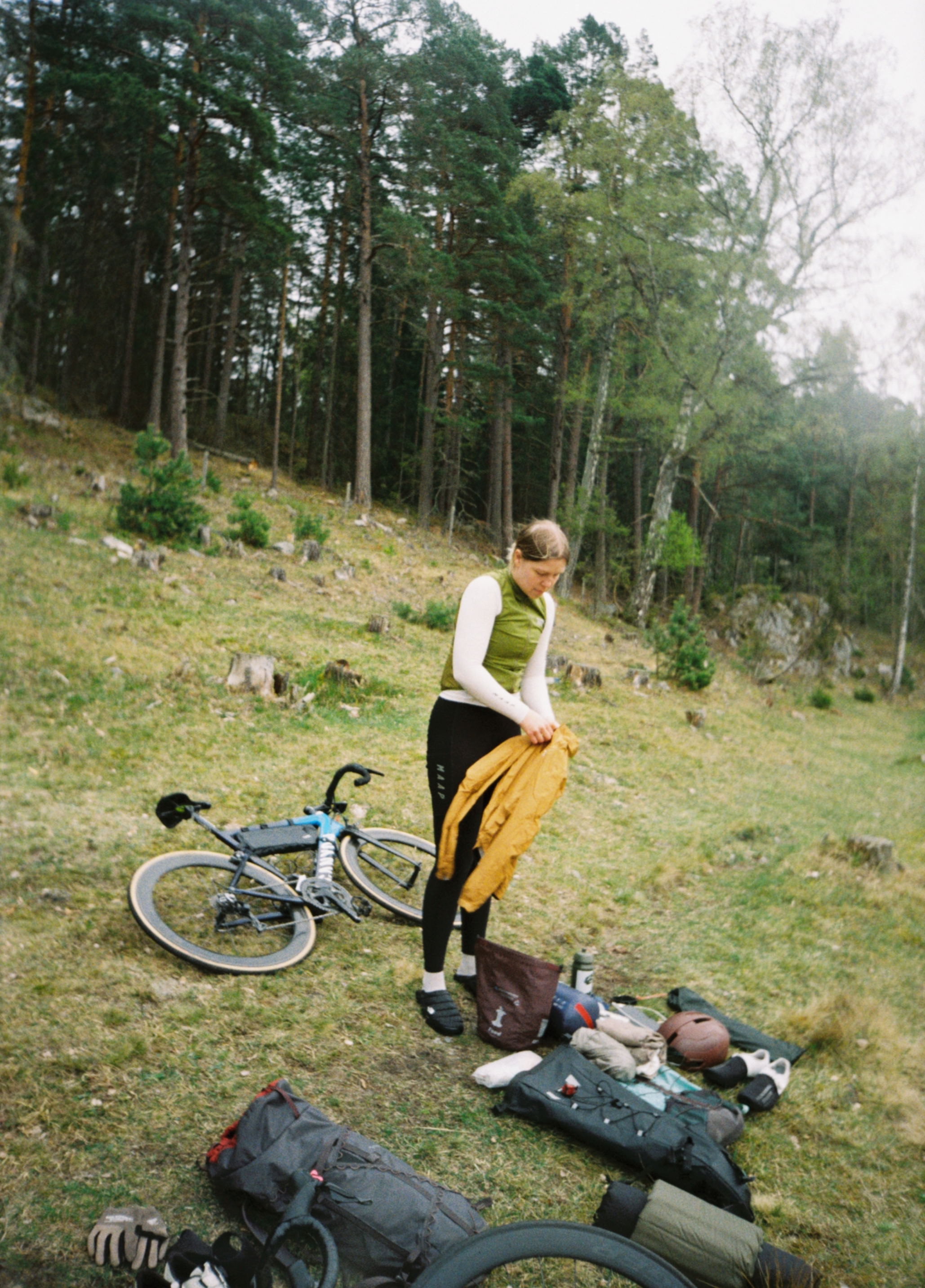 A woman standing outdoors in a forested area, packing or unpacking camping or biking gear with a bike lying on the ground nearby.