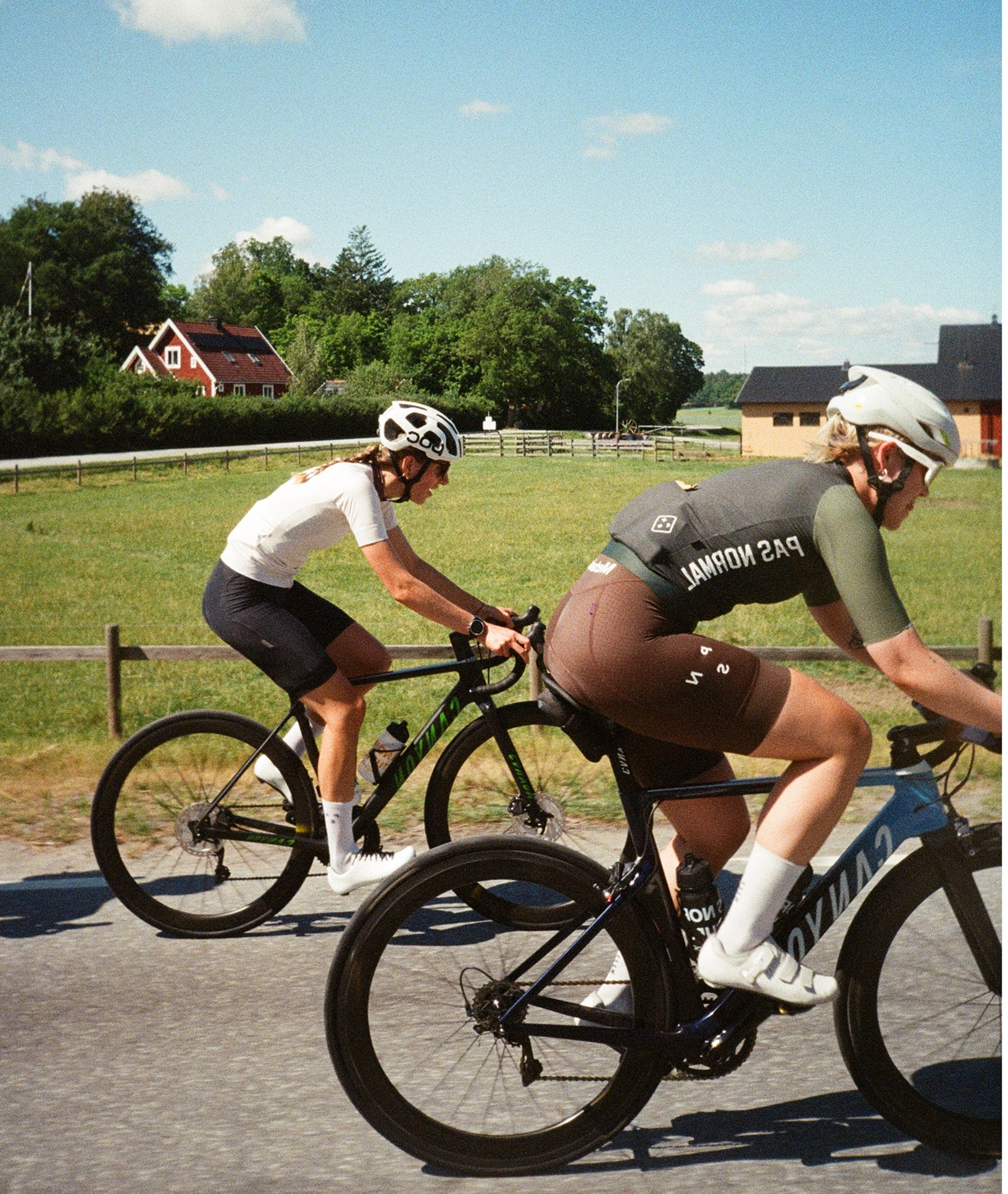 Two people riding bicycles on a paved road in a rural area under a clear blue sky, wearing helmets and athletic clothing.
