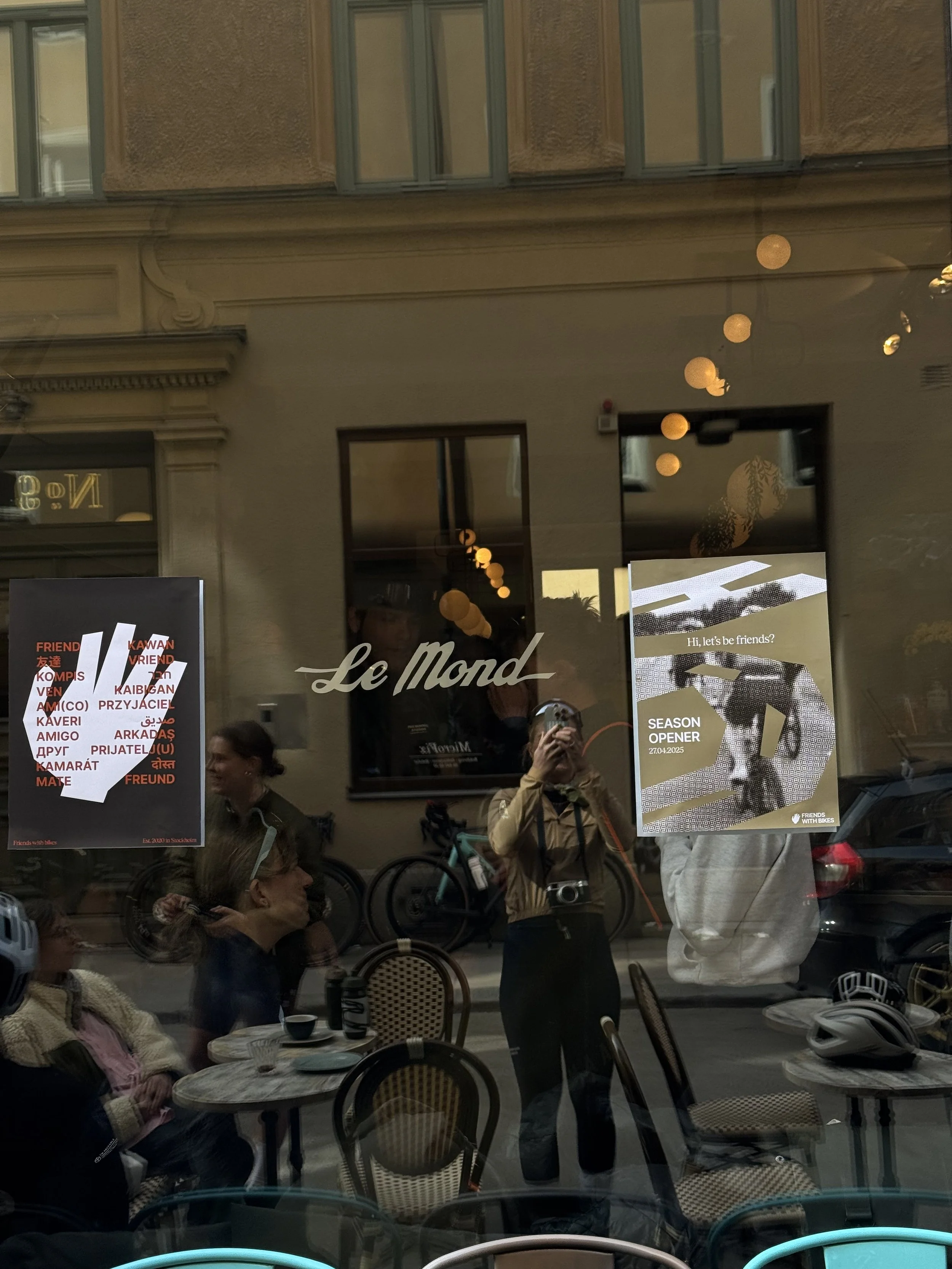 Reflection of people sitting inside a cafe, with posters on the window, including one for 'Le Monde' and another with a cyclist and the text 'Hi, let's be friends?'.