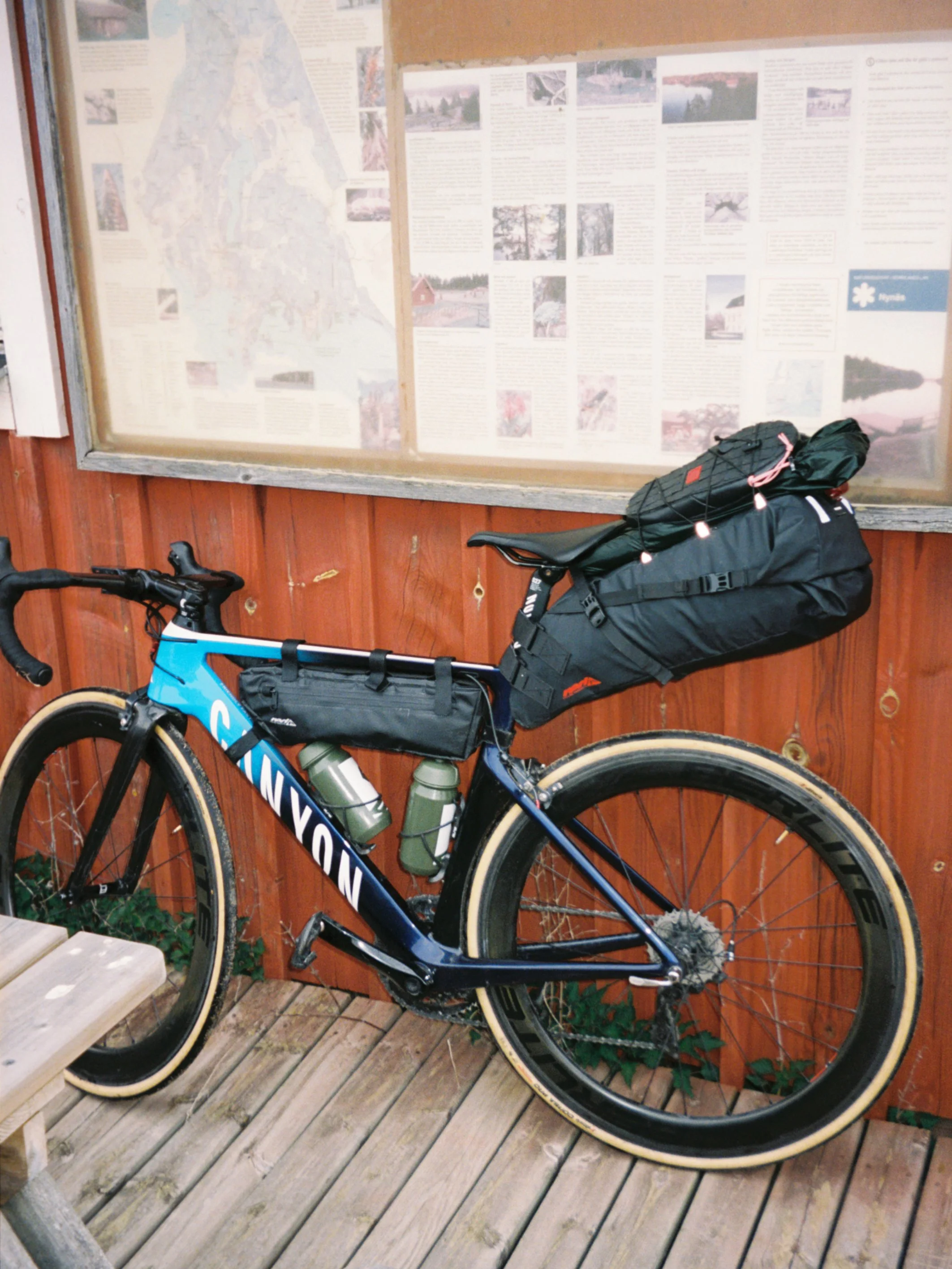 A blue Canyon mountain bike with beige tires equipped with two water bottles, resting against a wooden wall next to a large information display case with maps and text.