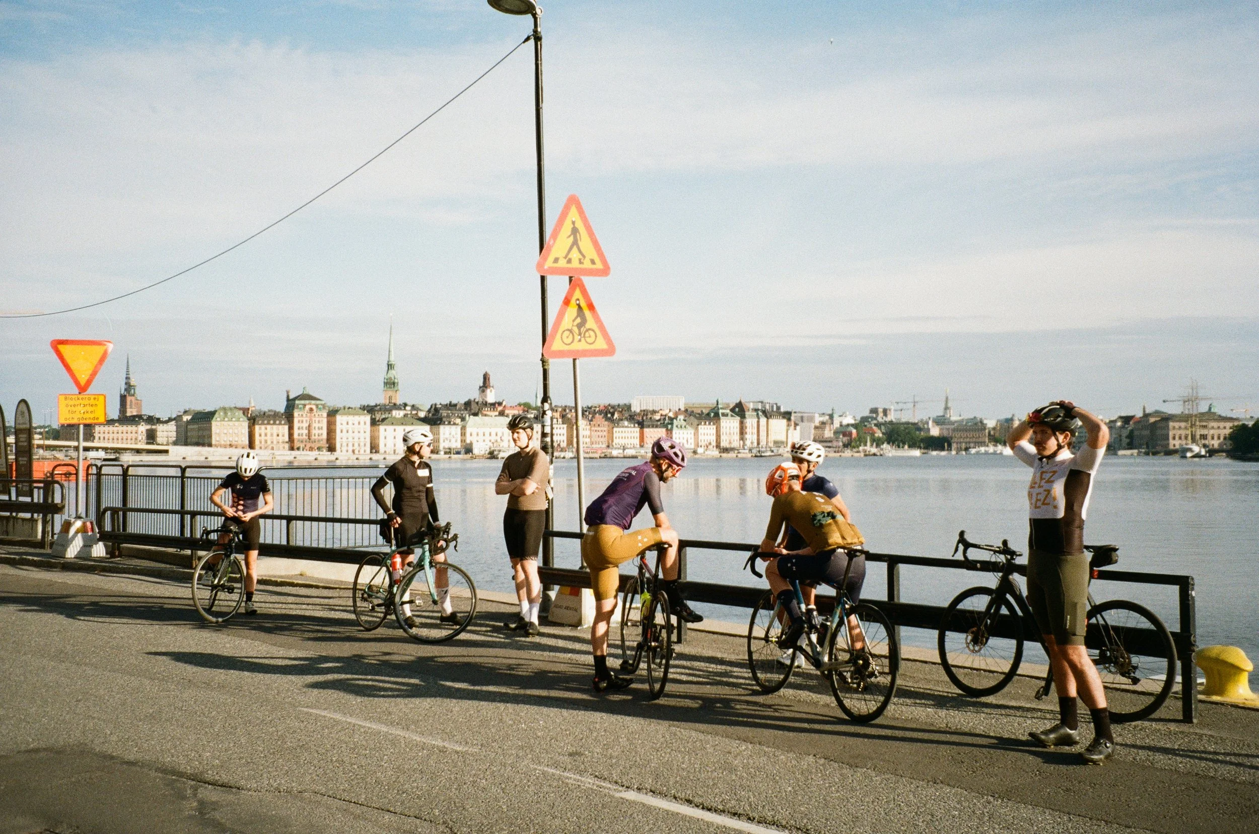 A group of cyclists and pedestrians on a bridge by a river with a city skyline in the background, some waiting and others preparing to ride.