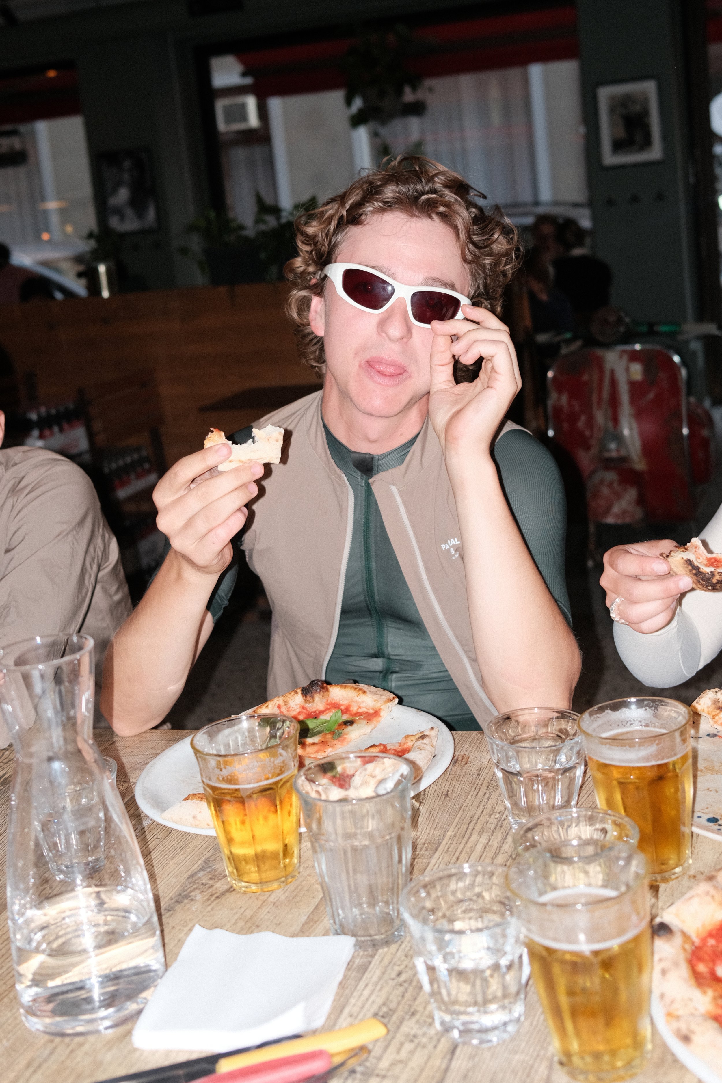 Two cyclists riding on a gravel road surrounded by greenery, with the cyclist in the foreground smiling and holding a water bottle.
