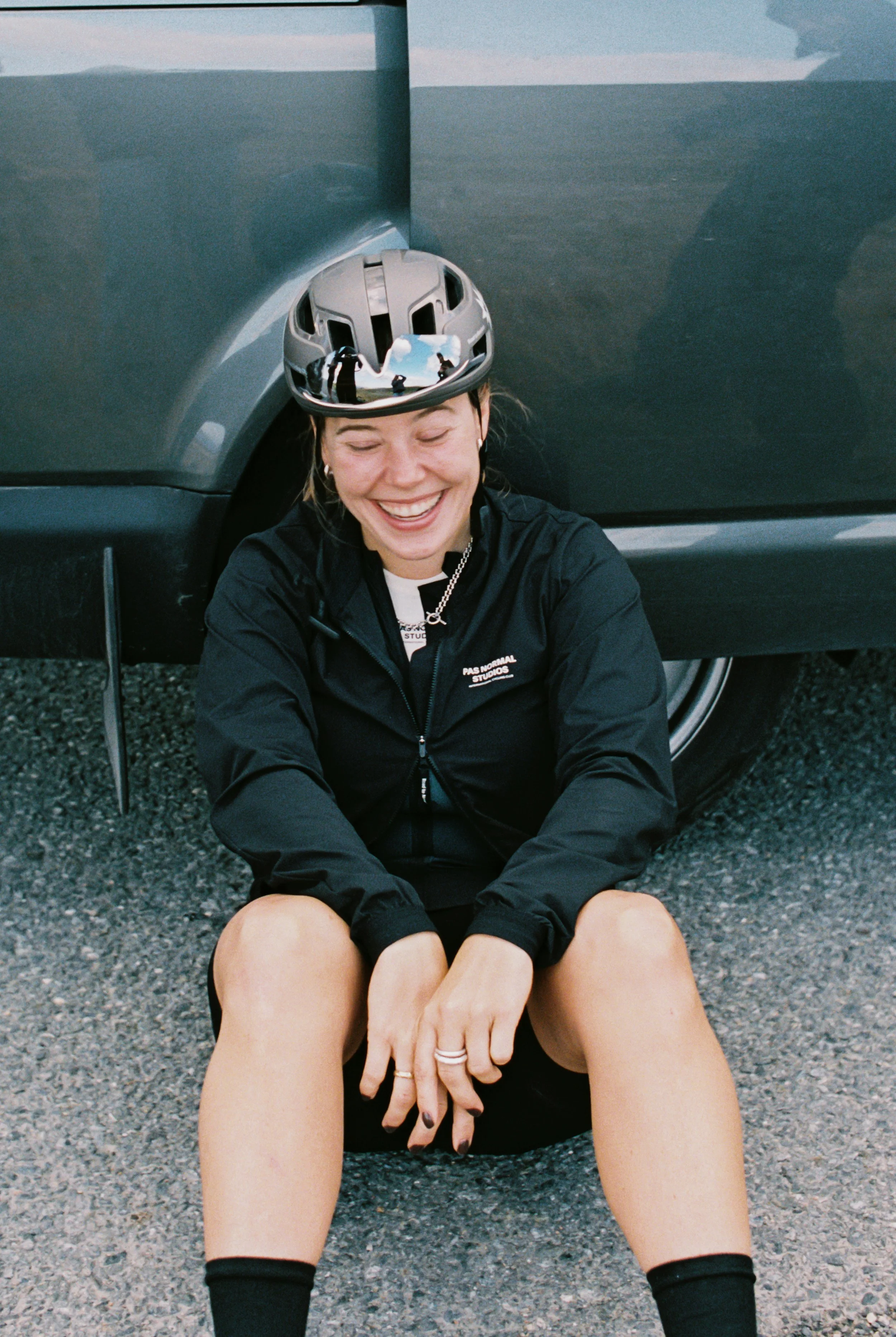 A woman sitting on the ground with her back leaning against a car, wearing a helmet and smiling.