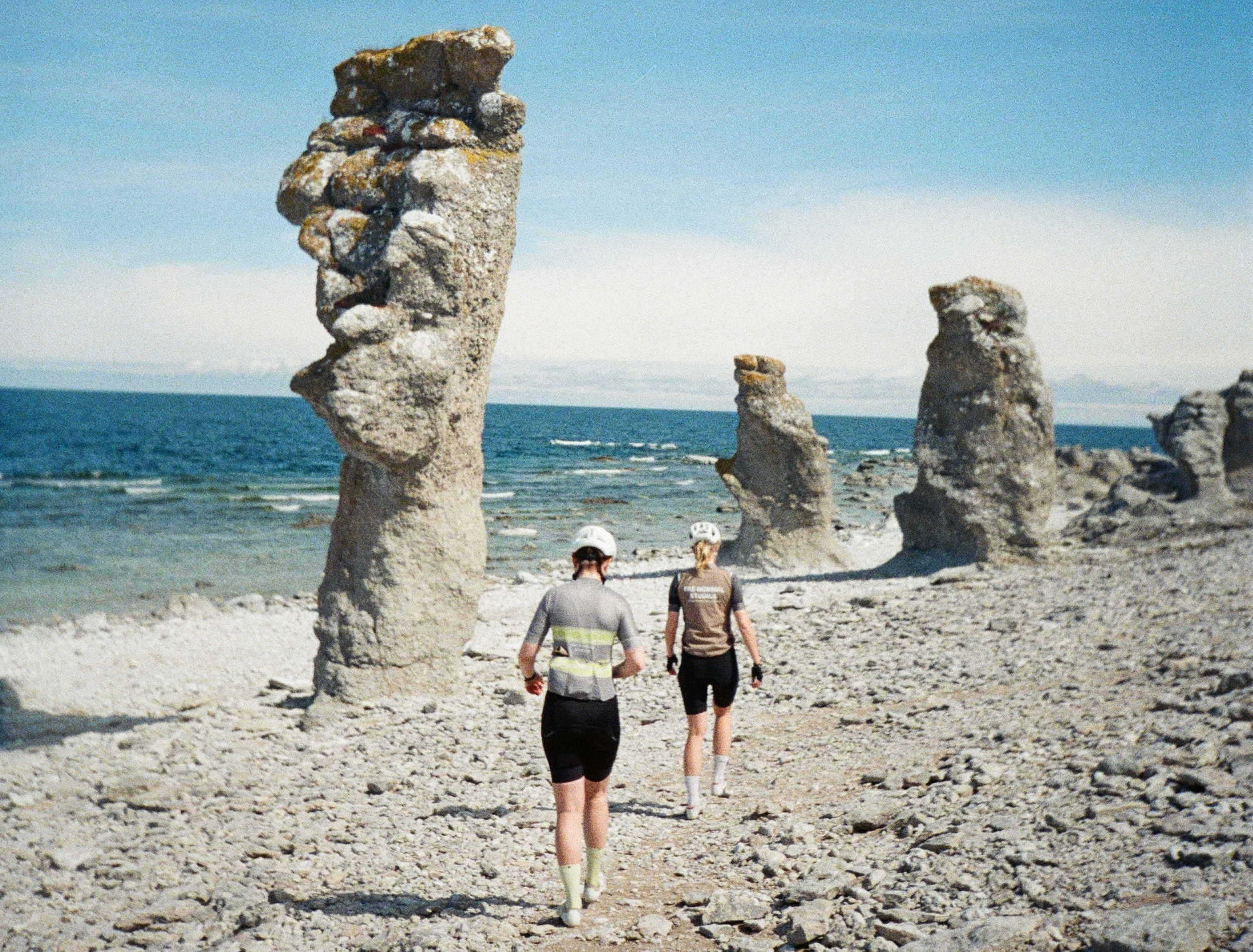 Two cyclists with helmets and cycling gear walking on a rocky beach with large limestone formations near the ocean under a partly cloudy sky.
