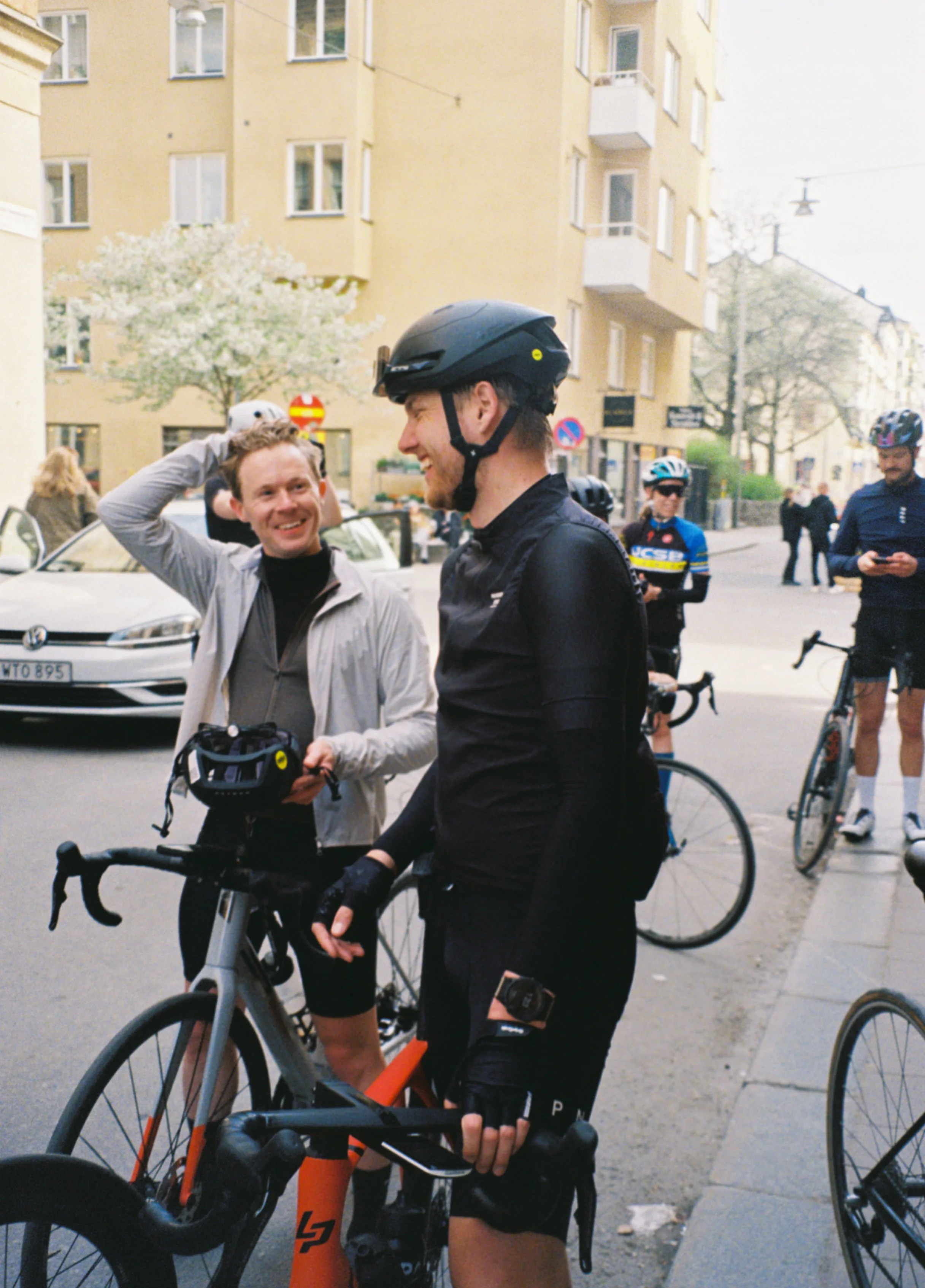 Two men with cycling gear and helmets standing on a city street, smiling and talking, with bicycles and other cyclists in the background.