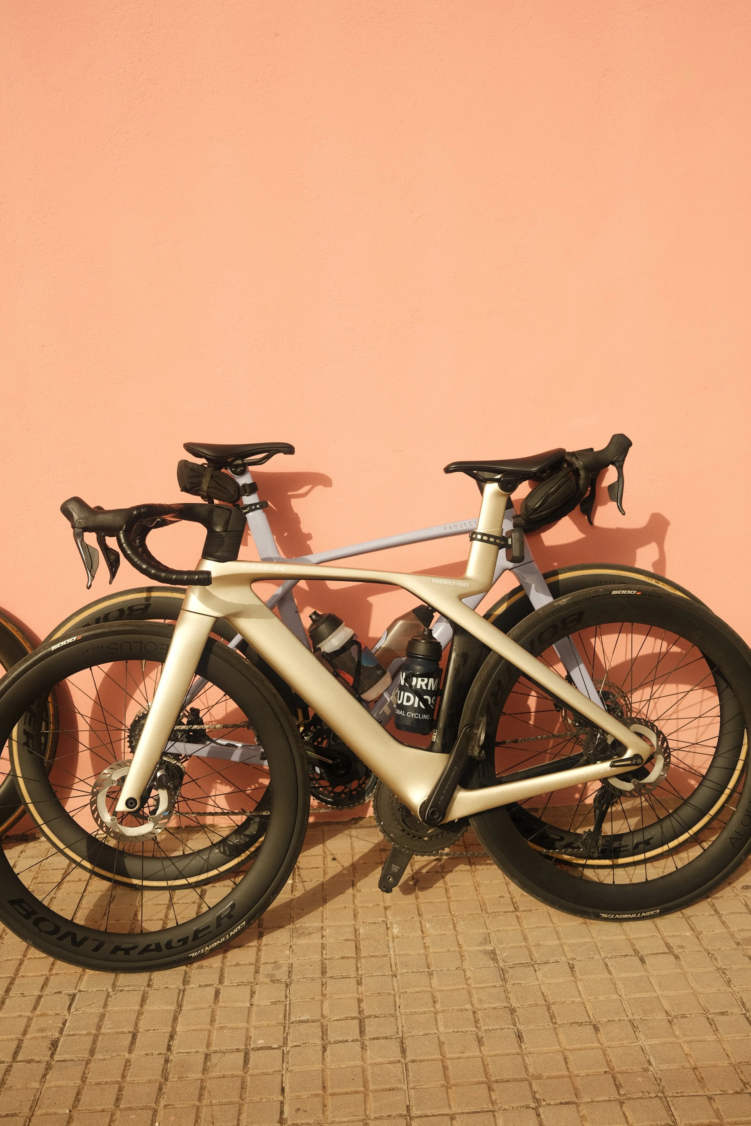 A beige and gray road bicycle with black accessories, resting against a peach-colored wall on a tiled floor.