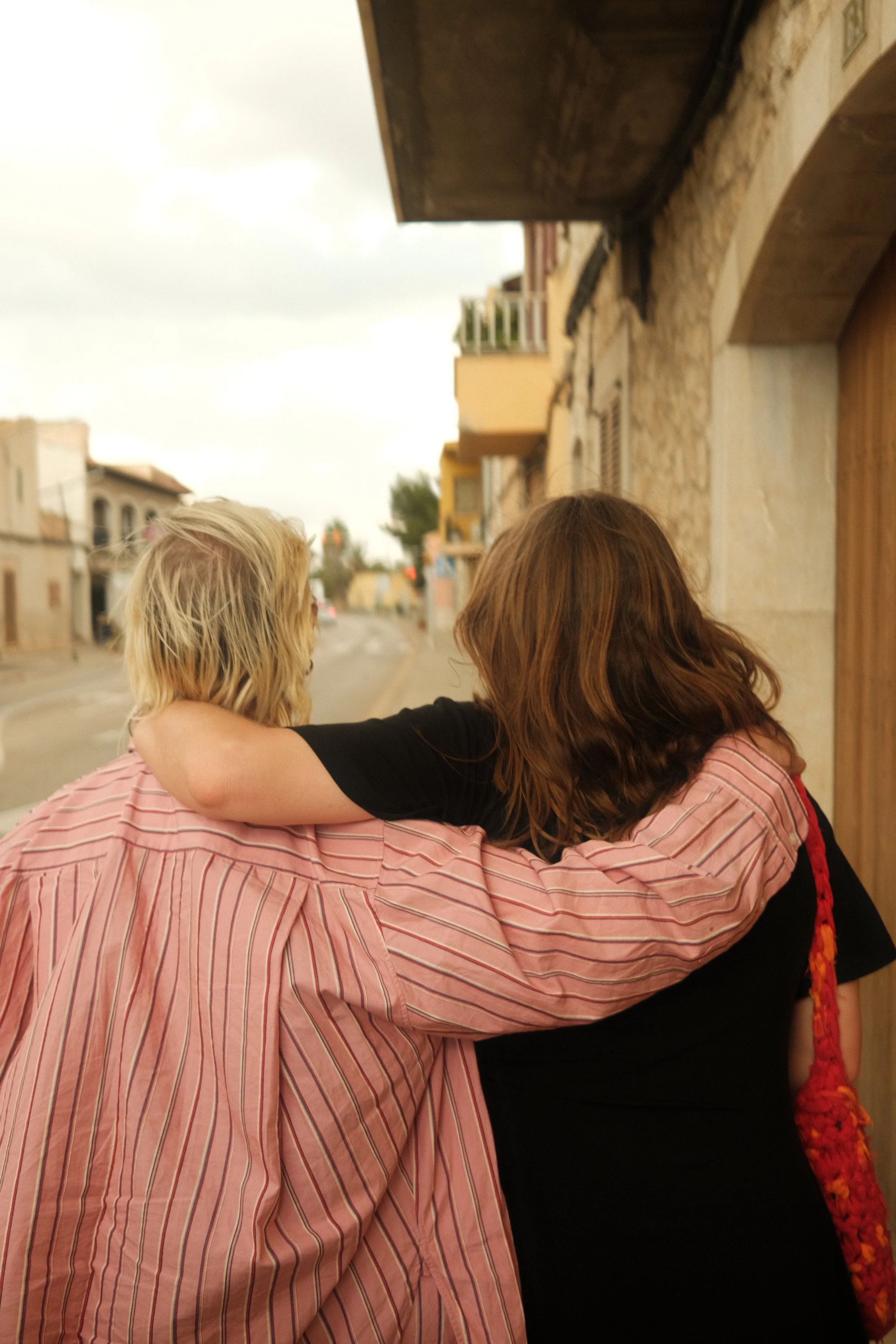 Two women with their backs to the camera, one with blonde hair and the other with brown hair, hugging each other on a street lined with buildings.