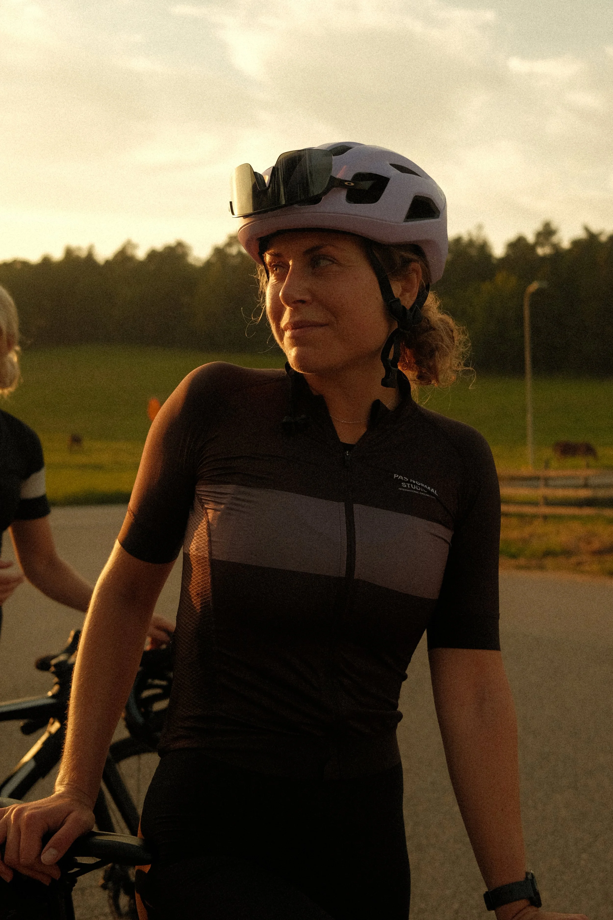 A woman wearing a white cycling helmet with sunglasses on top, dressed in a black and gray cycling jersey, standing outdoors during sunset, holding her bicycle handlebar, with another cyclist partially visible in the background.
