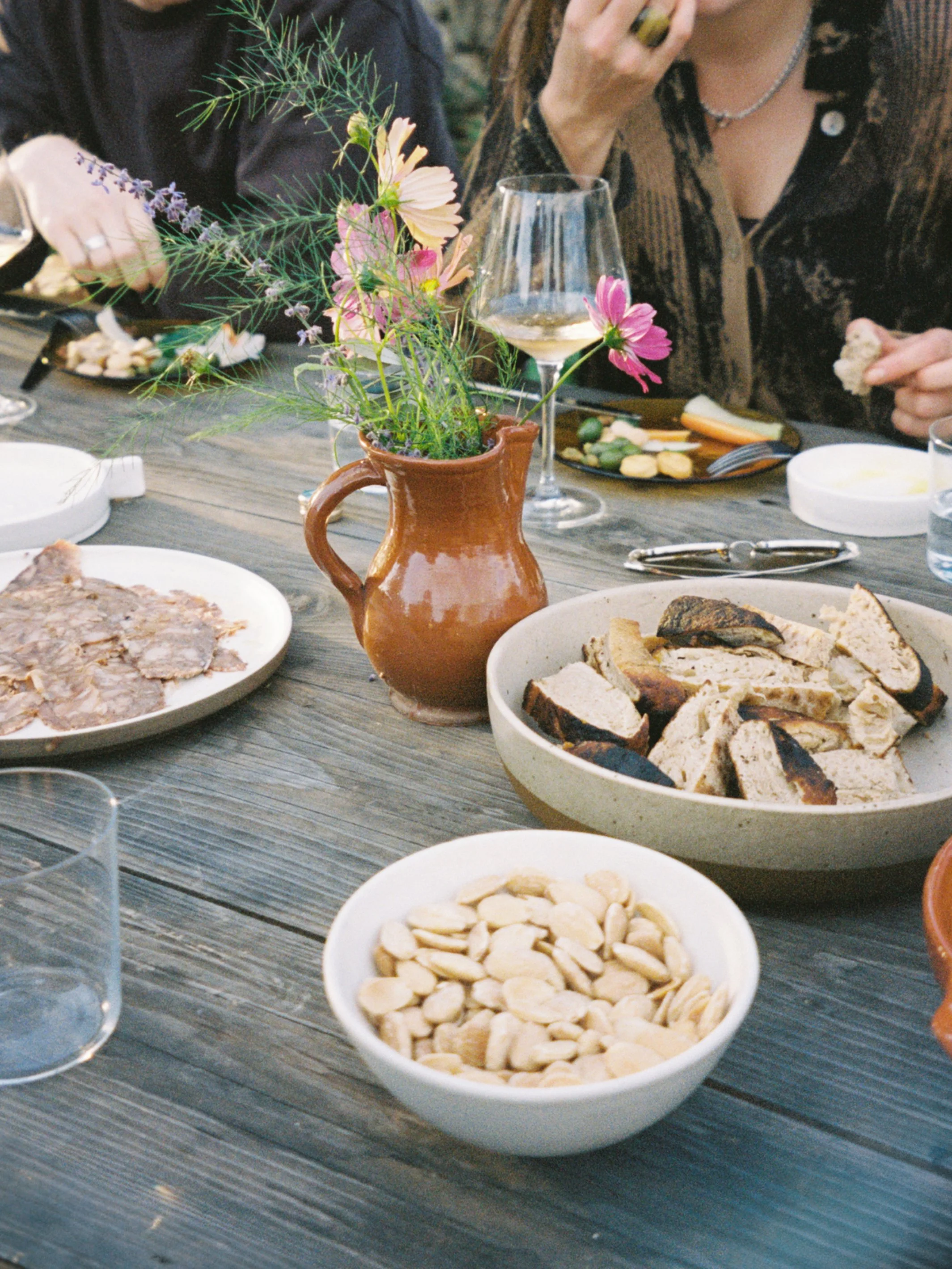 A rustic outdoor dining table with a terracotta pitcher of wildflowers, bowls of sliced bread, cashews, and baked dishes, and guests enjoying wine and food.
