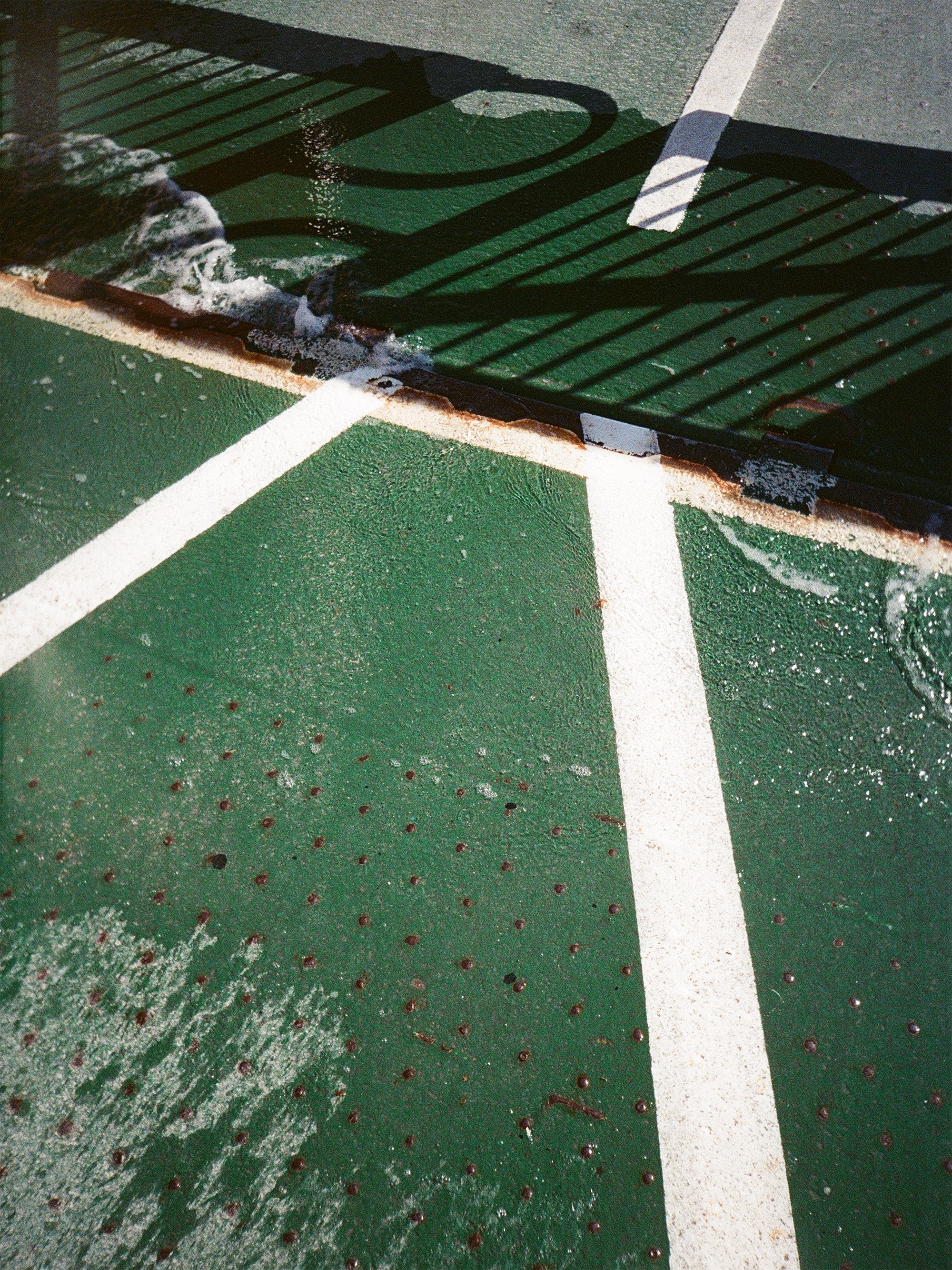 Section of a green outdoor tennis court with white painted lines, showing water pooling and a shadow of a fence.
