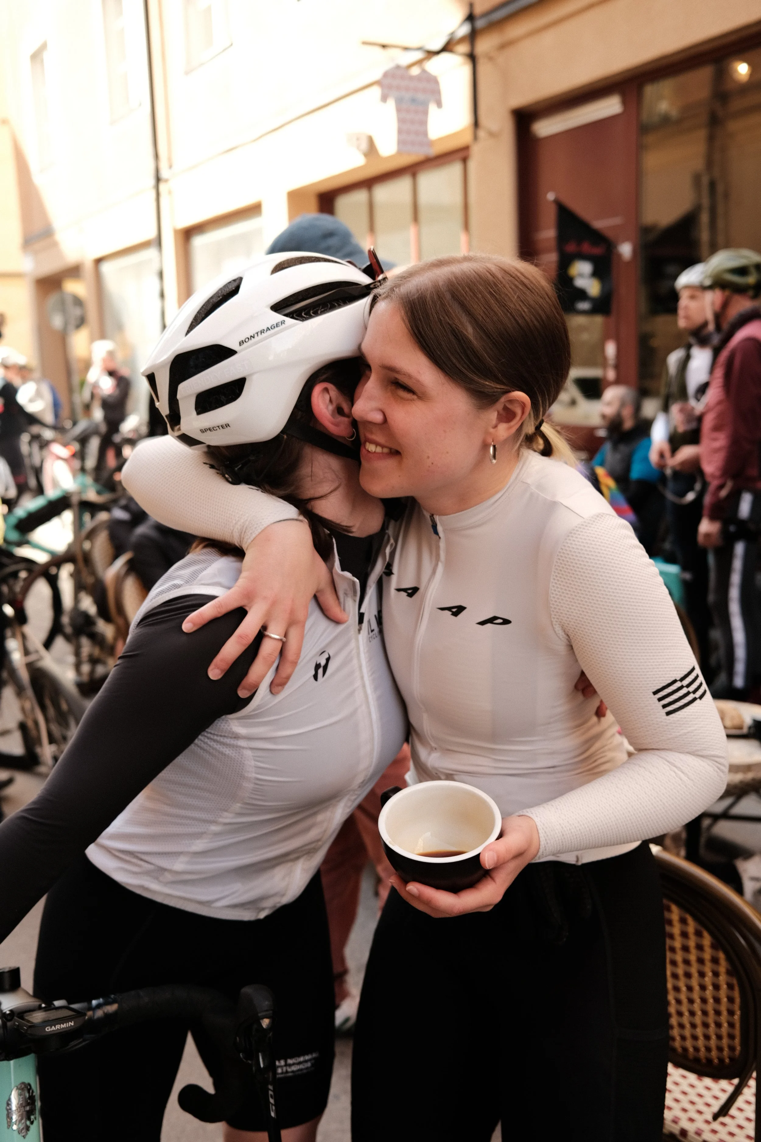 Two women embracing in a street, one wearing a bicycle helmet and cycling attire, the other holding a cup and smiling, with a crowd and bicycles in the background.