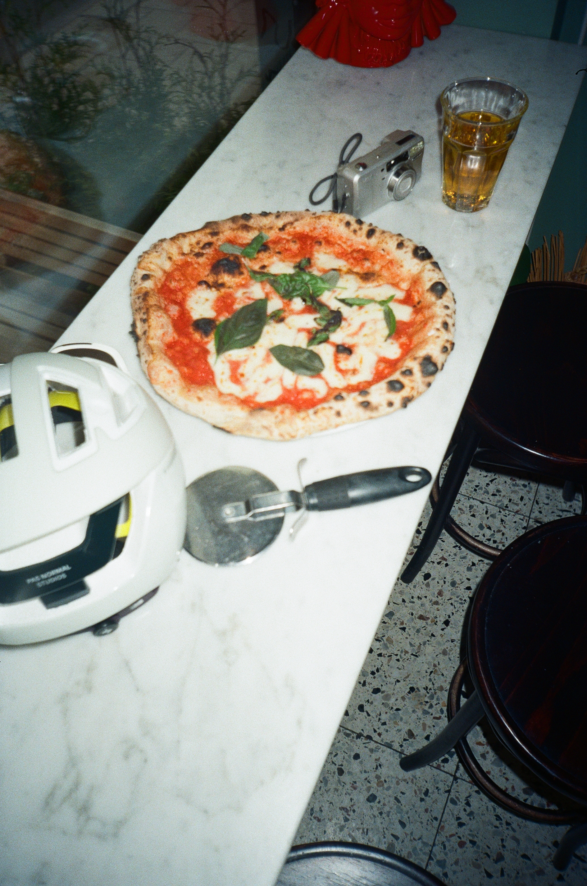 A Margherita pizza with basil leaves on a marble countertop, with a camera, a glass of beer, a pizza cutter, and a white helmet nearby.