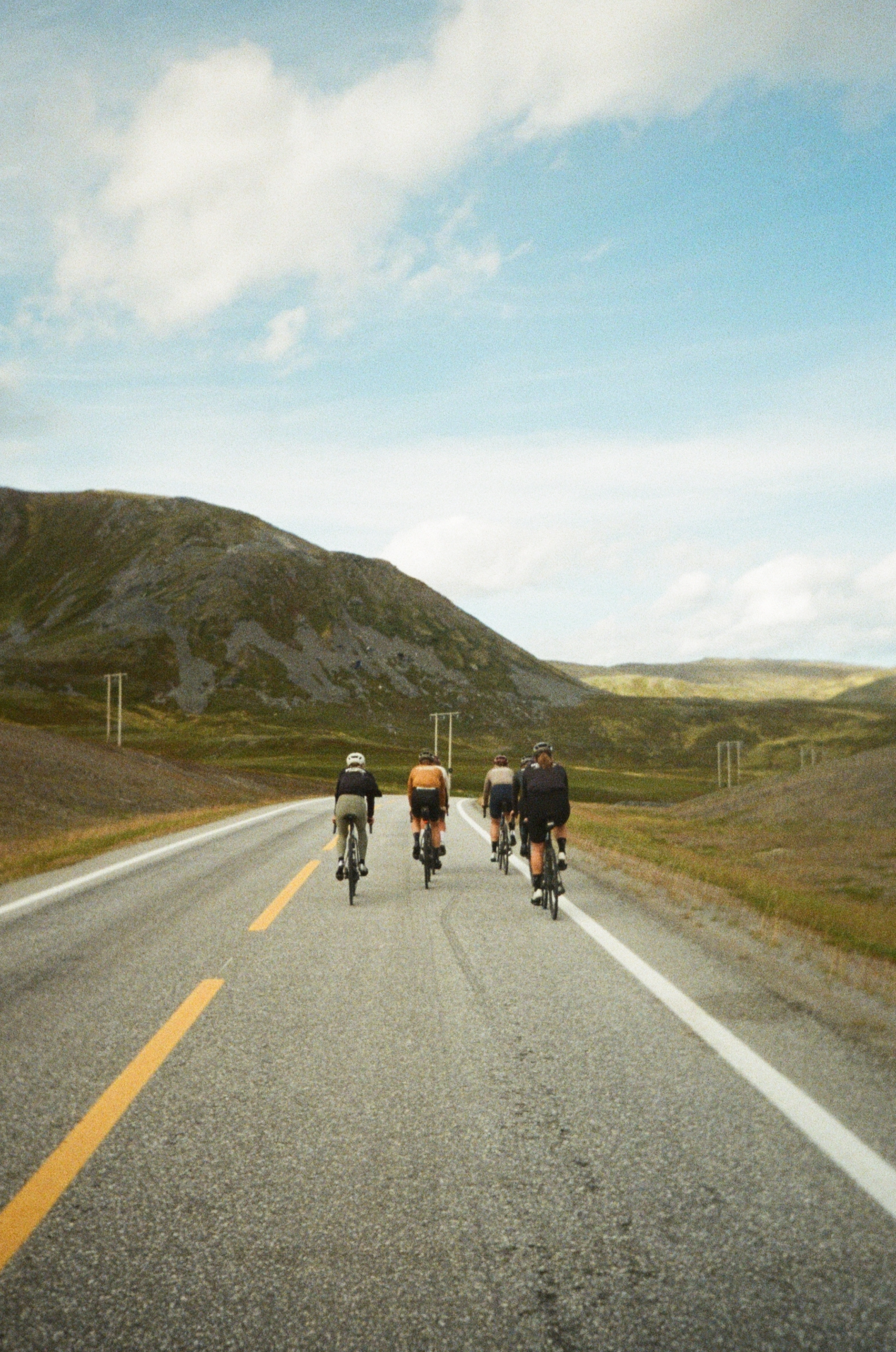 A group of cyclists riding on a two-lane road through a hilly landscape with a partly cloudy sky.