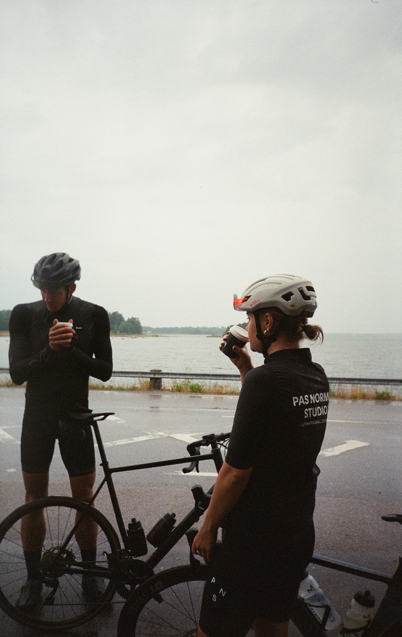 Two cyclists in black outfits and helmets stand near a body of water, one drinking coffee and the other looking at a phone.