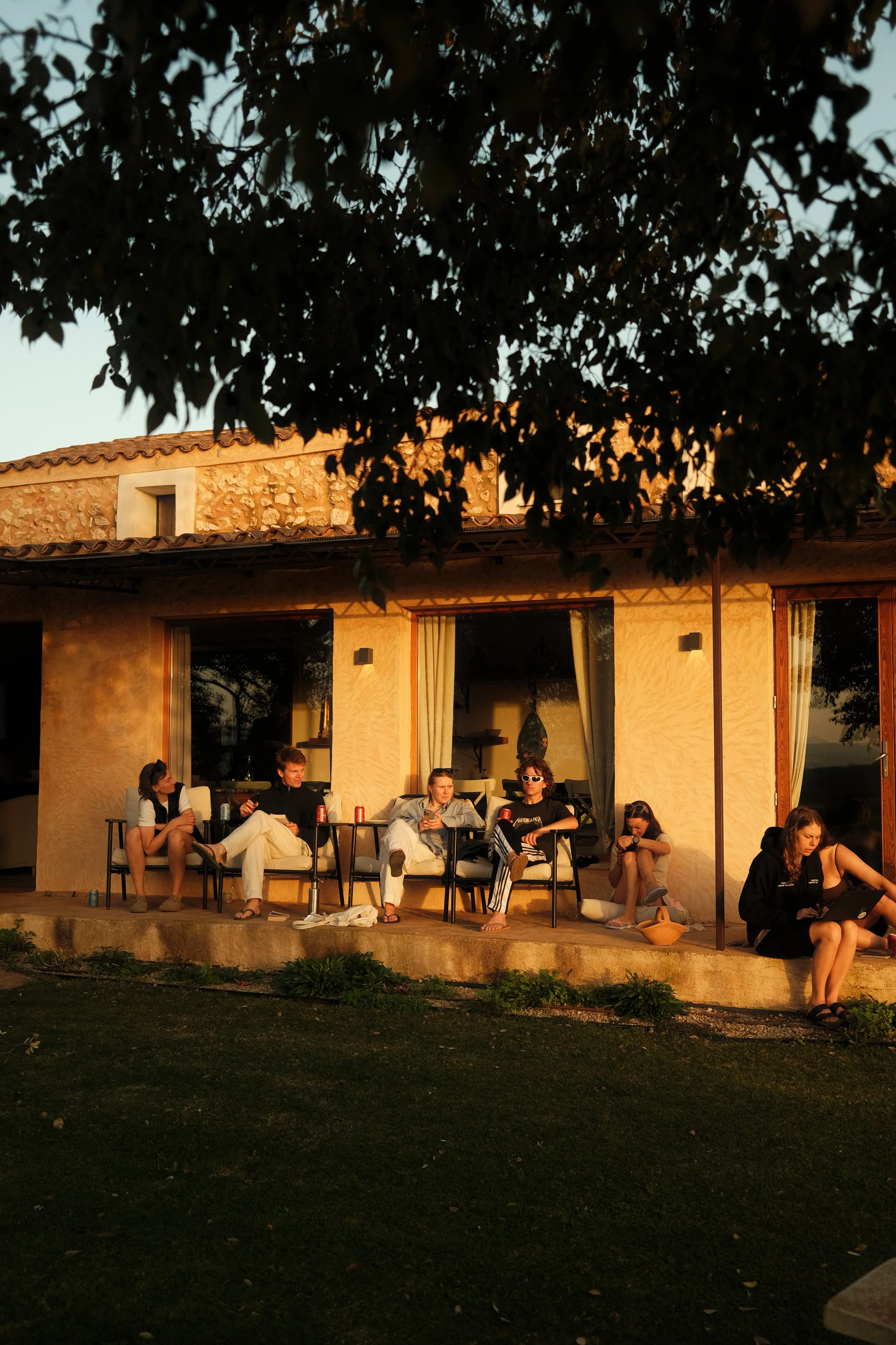 A group of six young people sitting on chairs and the ground outside a house, with some drinking soda and using phones, during sunset.